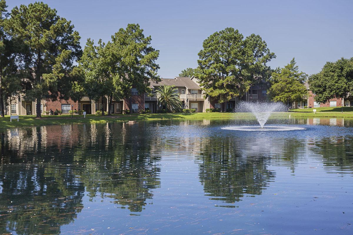 A tranquil pond surrounded by lush greenery and residential buildings. A fountain sprays water in the center of the pond, creating ripples on the surface. Clear blue sky reflects in the water, enhancing the peaceful atmosphere of the scene.