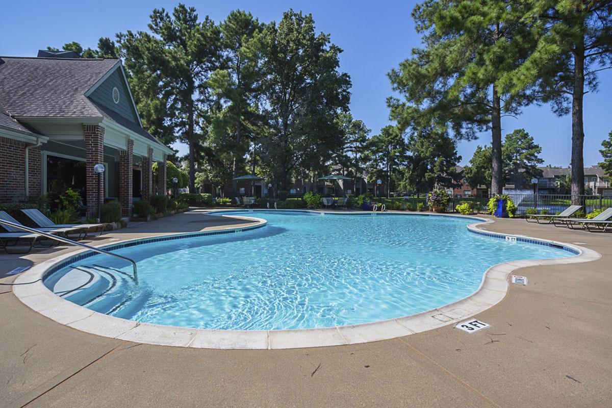 A clear, inviting swimming pool surrounded by lounge chairs and trees. The pool has a curved shape and is set against a bright blue sky. Nearby, there is a clubhouse with brick exterior, adding to the tranquil outdoor atmosphere. The area is well-maintained, perfect for relaxation and recreation.