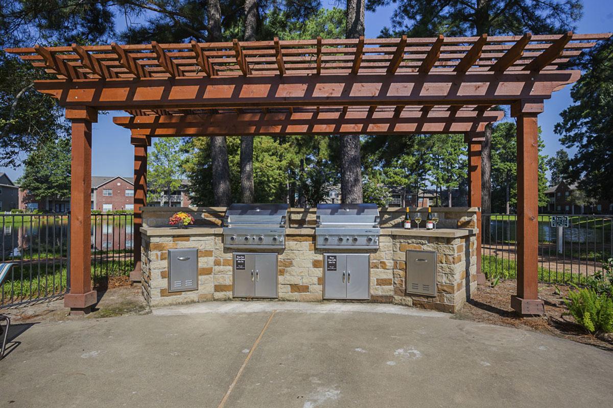 Outdoor barbecue area featuring three built-in grills under a wooden pergola. The setup includes stone accents, a flower planter, and a concrete pad. Lush green trees and a fenced area in the background create a serene outdoor atmosphere, ideal for gatherings and cooking.