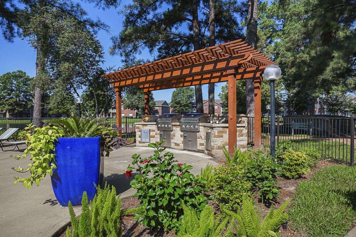 An outdoor grilling area features a stone barbecue with a wooden pergola overhead. Surrounding the grill are vibrant green plants and flowers, including a large blue planter. The setting is bright and sunny, with trees and a fence in the background, creating a pleasant courtyard atmosphere for social gatherings.