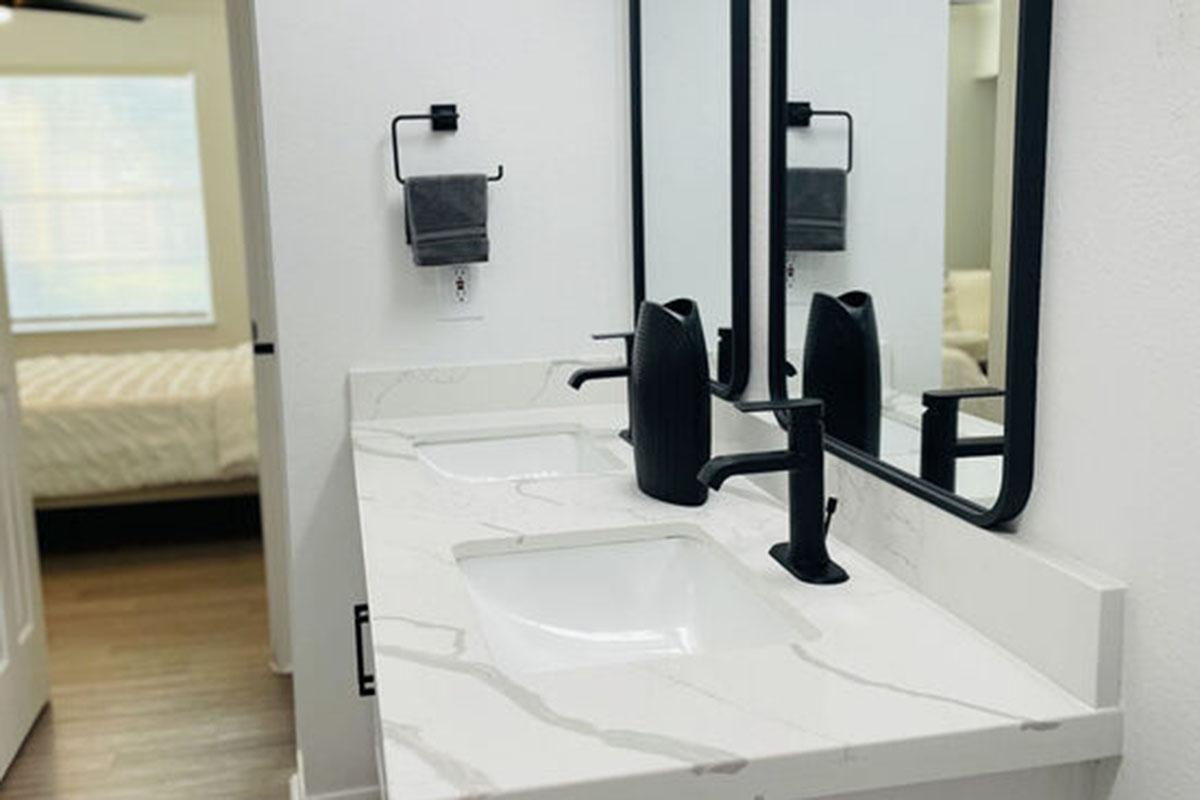 A modern bathroom featuring a double vanity with a marble countertop, two rectangular white sinks, and sleek black faucets. Above each sink, there are large circular mirrors. A grey towel is hanging on a holder nearby, and a softly lit bedroom is visible through an open door in the background.