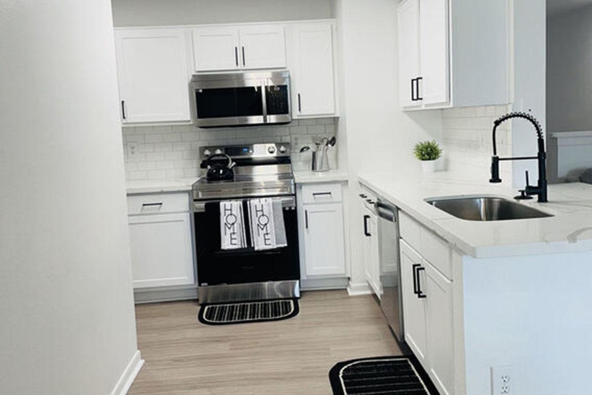 Modern kitchen with white cabinets, stainless steel appliances, and a black faucet. A decorative plant sits on the countertop. Two towels with "HOME" printed on them hang by the sink. The flooring is light wood, and there are black and white mats on the floor, creating a clean and inviting space.