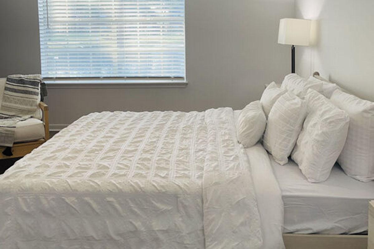 A neatly made bed with white bedding and several fluffy pillows in a light-colored bedroom. There's a comfortable chair in the corner and a lamp beside the bed. Natural light filters through a window with blinds, creating a calm and inviting atmosphere.