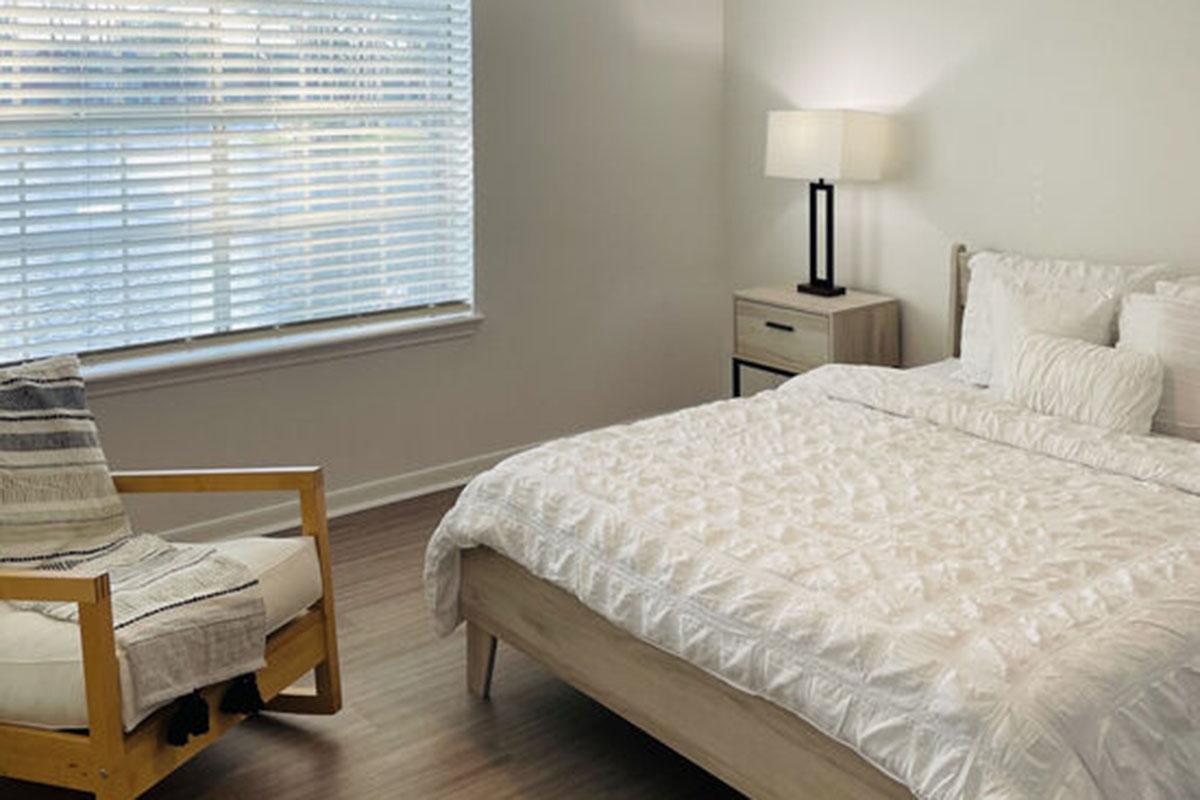 A bright and modern bedroom featuring a neatly made bed with white bedding, a wooden bed frame, and a side table with a lamp. A comfortable chair with a throw blanket is positioned near a window with white blinds, allowing natural light to fill the room. The flooring is a light wooden finish, contributing to a serene atmosphere.
