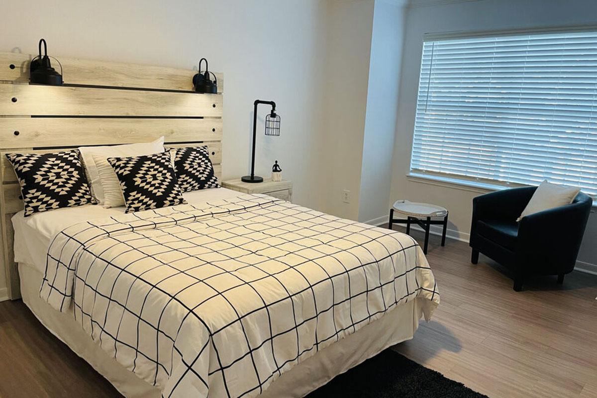 A modern bedroom featuring a light wood bed frame with a grid-patterned bedding, two black and white patterned pillows, a nightstand with a lamp, and a black armchair next to the bed. Natural light filters through a window with blinds, illuminating the neutral-colored walls and flooring.