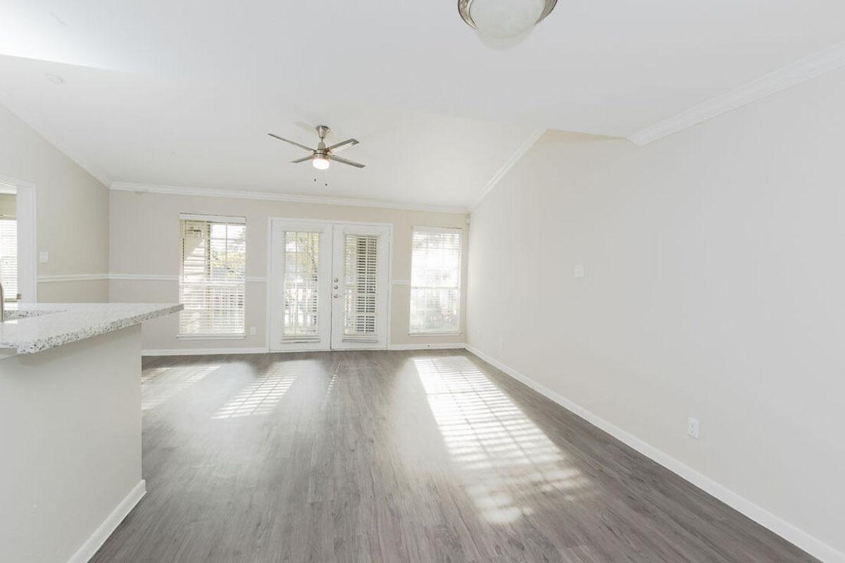 Spacious living area with light-colored walls and laminate flooring. Features include a ceiling fan, a bar counter, and large windows that let in natural light. The room opens to a patio with sliding glass doors and has a cozy, airy feel.