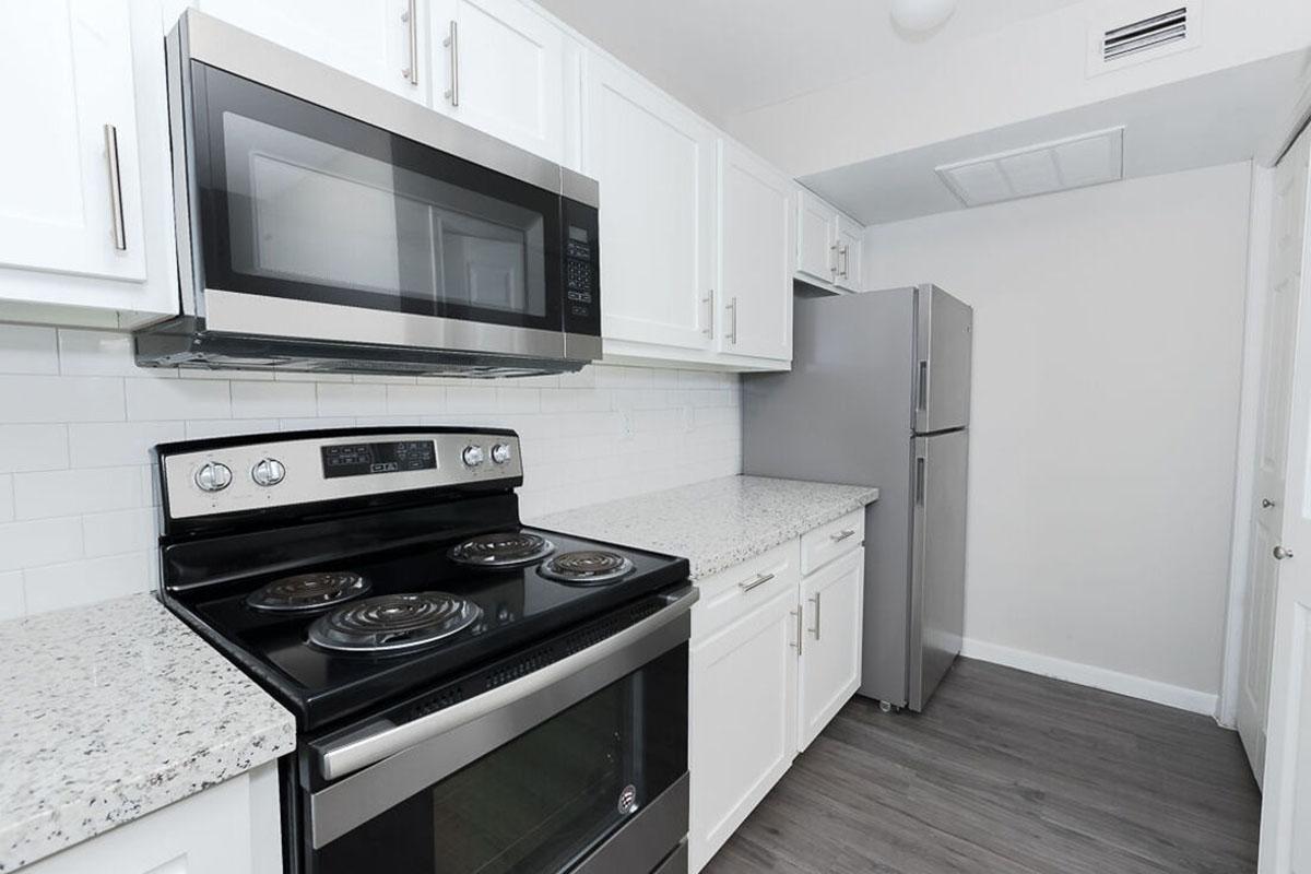 Modern kitchen featuring white cabinetry, a stainless steel microwave above a black stove, and a silver refrigerator. The countertops are speckled gray and white, and the flooring is a light-colored laminate. The walls are painted in a neutral tone, creating a bright and clean appearance.