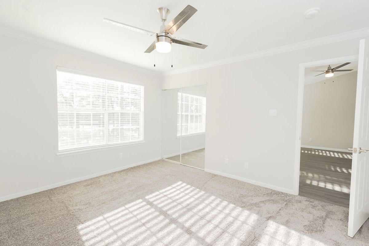 A spacious, well-lit bedroom featuring light-colored walls and carpet. There is a ceiling fan with five blades and a large window with drawn blinds allowing sunlight to filter in. A mirrored closet door is visible on one wall, complementing the neutral decor and bright atmosphere of the room.