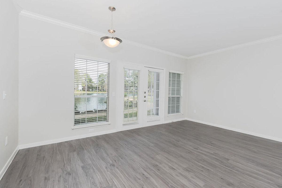 Empty living room with light gray walls and wood-style flooring. Contains large windows with white blinds, allowing natural light in, and a sliding glass door that opens to a view of a lake. A modern ceiling light fixture hangs from the ceiling. The space is unfurnished and well-lit.
