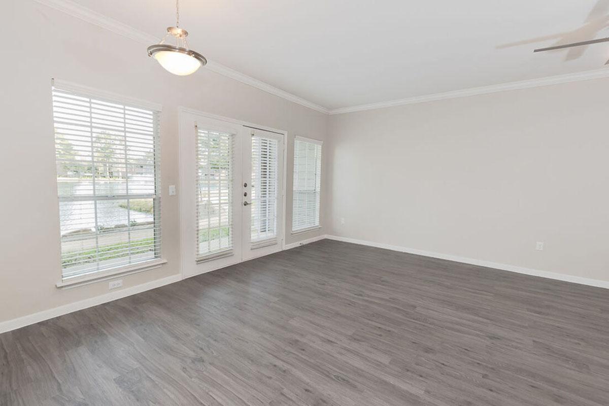 A bright, empty living room featuring light gray walls, large windows with blinds, and a ceiling fan. The floor is covered with sleek, dark laminate flooring, and there is a set of double doors leading outside. The room is well-lit with natural light.