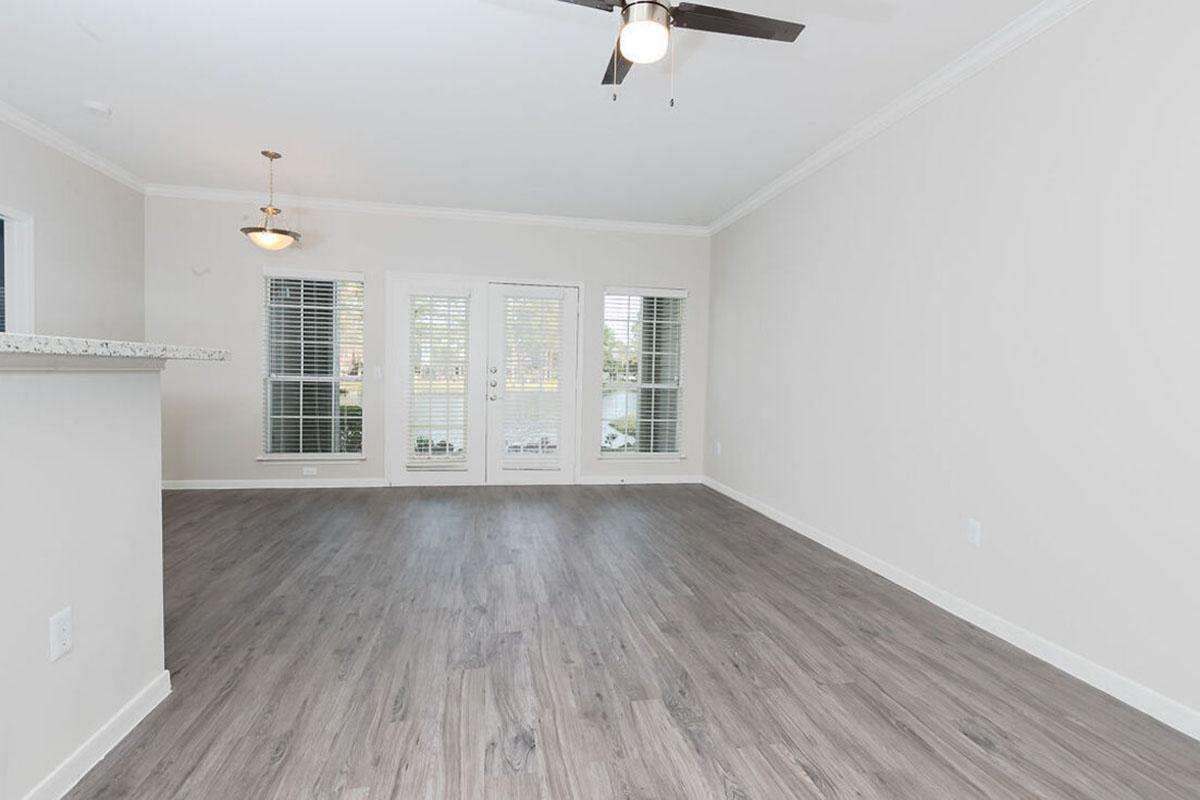 A spacious, light-filled living area with wood-look flooring, featuring a ceiling fan and large windows. There are glass doors leading outside, framed by vertical blinds. The walls are painted a soft neutral color, enhancing the bright and airy feel of the space.