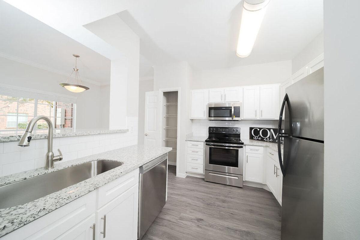 Bright, modern kitchen featuring white cabinetry, stainless steel appliances, a granite countertop, and a double sink. The open layout includes a large window allowing natural light and a view of the outside. Light-colored wood-style flooring complements the design.