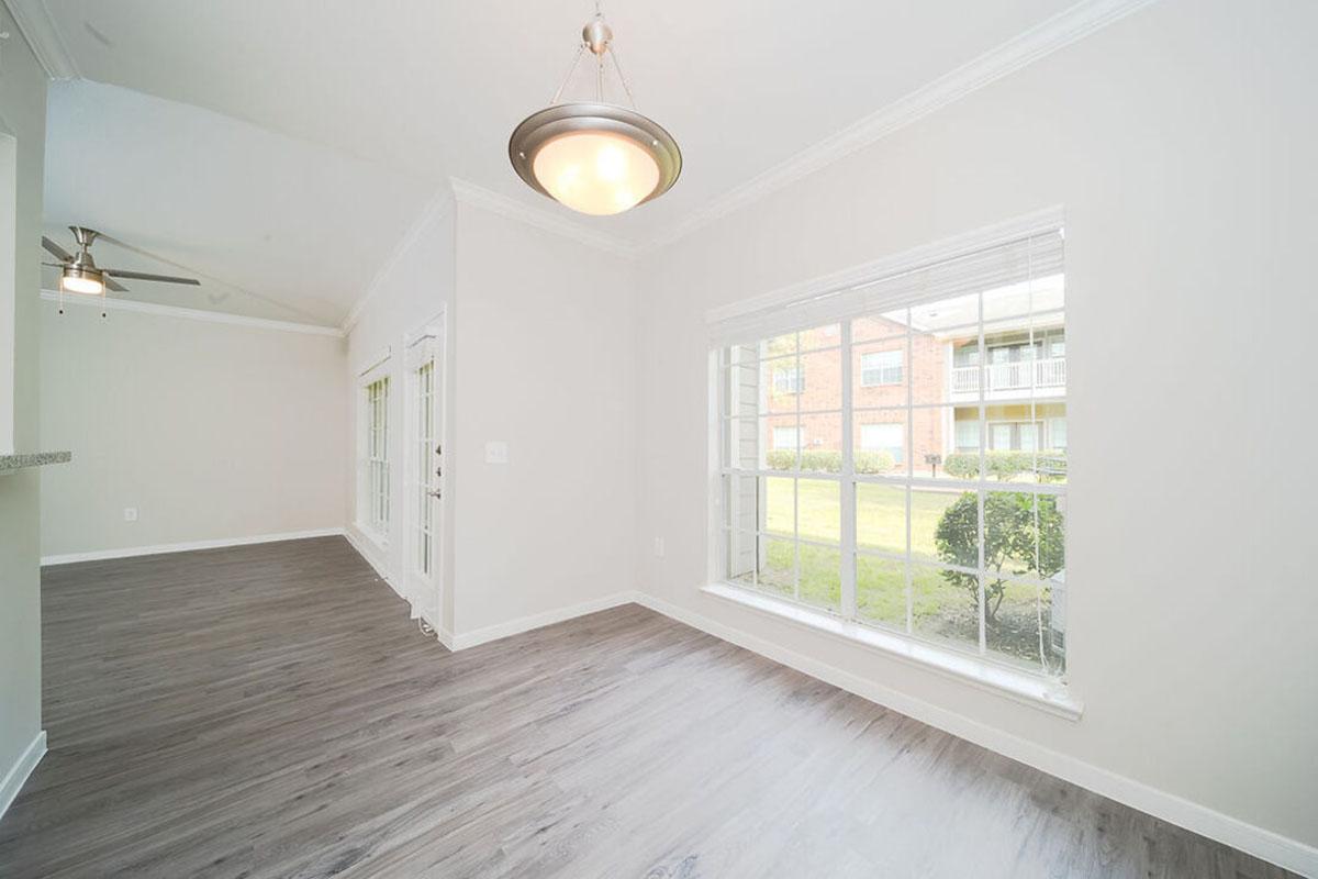 Bright and airy interior of a living space featuring a large window with views of outdoor greenery, light hardwood flooring, and neutral-colored walls. A modern ceiling light fixture is visible, along with a ceiling fan. The room appears open and inviting, creating a comfortable atmosphere.