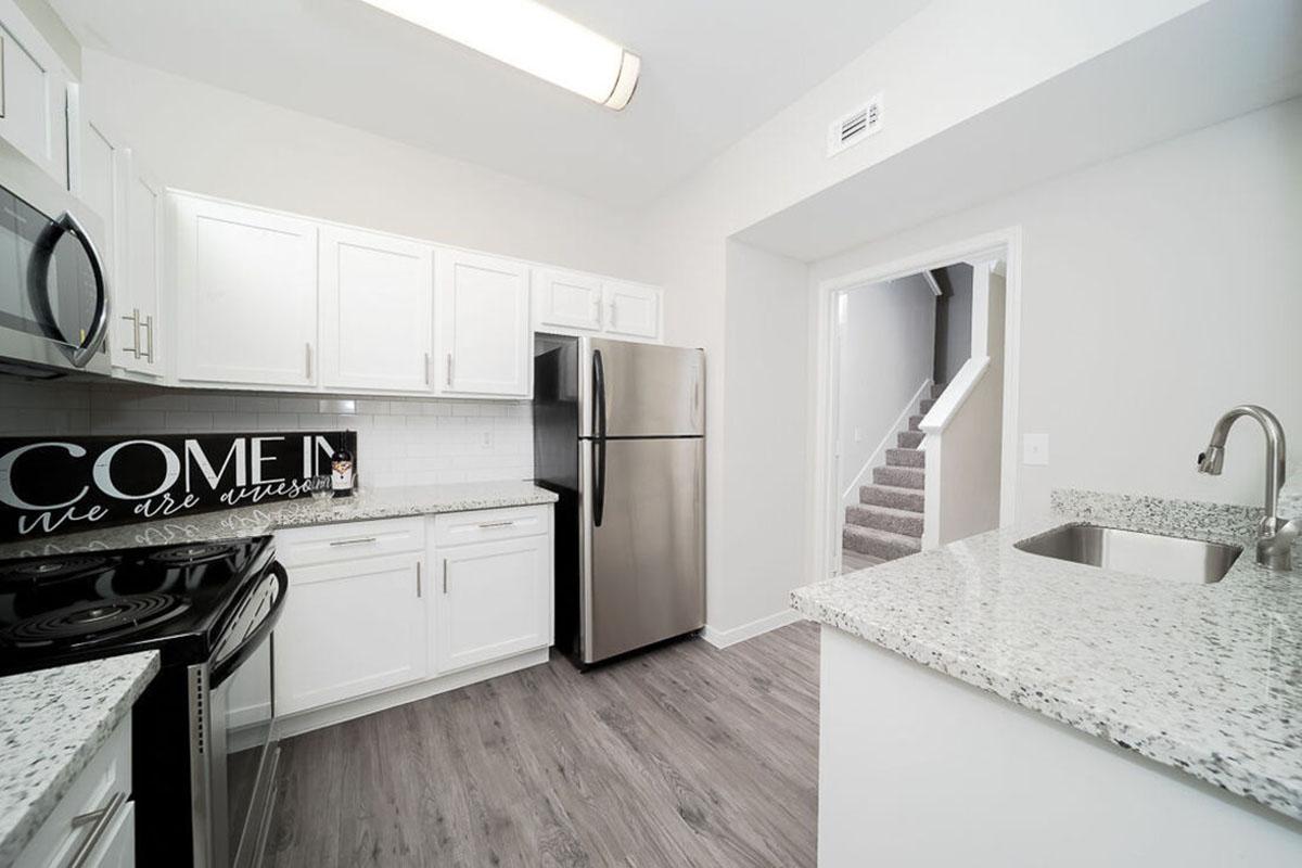 Modern kitchen featuring white cabinetry, stainless steel appliances, and speckled granite countertops. A decorative sign that reads "COME IN we are open" is displayed on the wall. There is a staircase visible in the background, and natural light is streaming from a ceiling fixture.