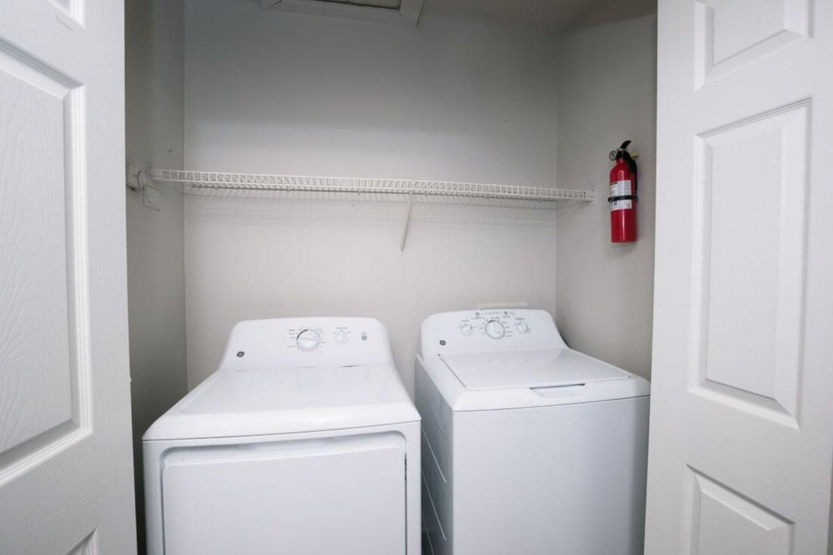 A laundry room featuring a stacked washer and dryer. The appliances are white and positioned side by side. Above them is a wire shelf for storage, and a fire extinguisher is mounted on the wall nearby. The room is well-lit with white walls and doors partially closed.