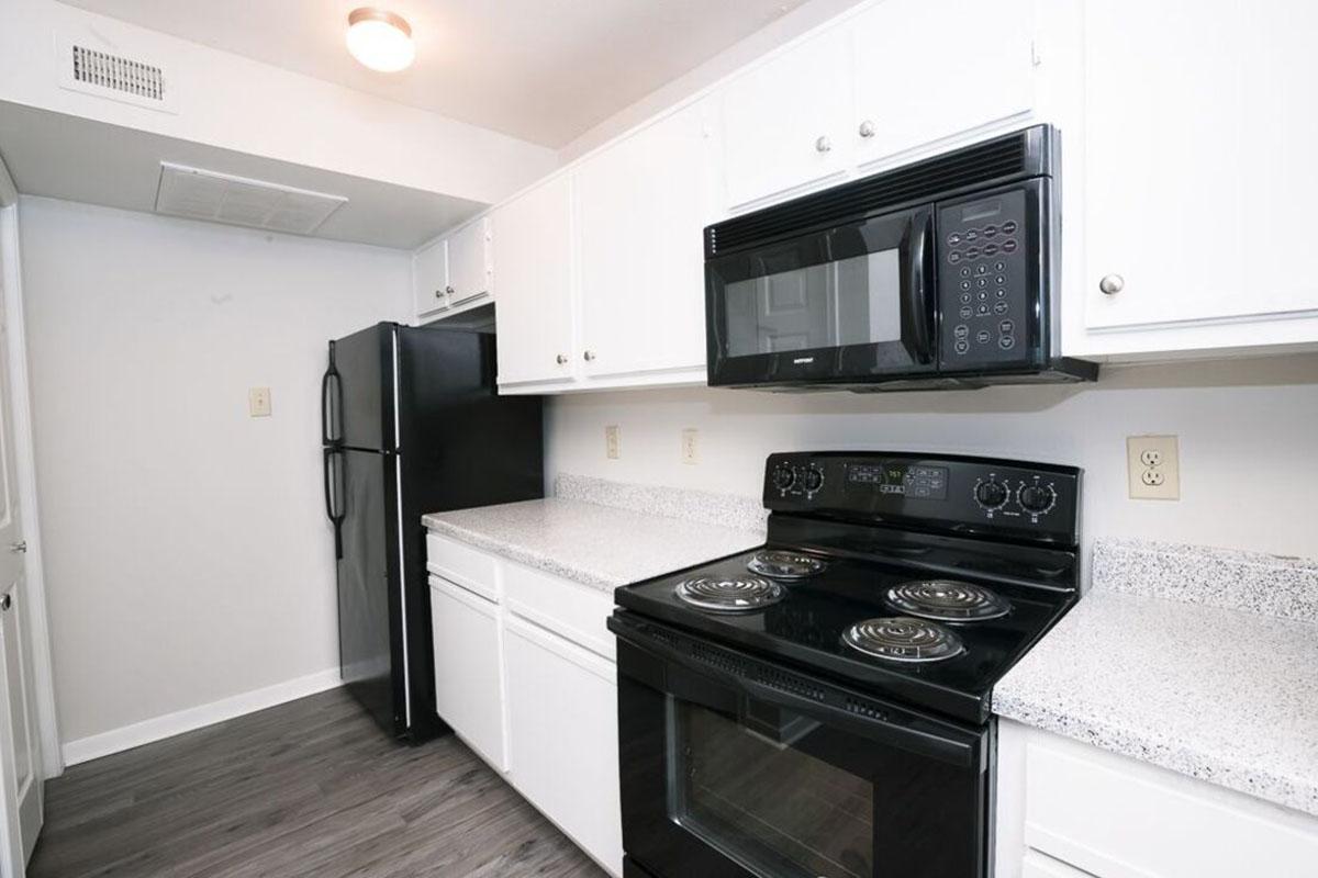 A modern kitchen featuring white cabinets, a black refrigerator, and a black stove with an oven. The countertop is light-colored, and there is a microwave above the stove. The flooring is a dark laminate. The space is well-lit with a ceiling light.