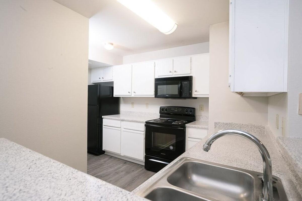 A modern kitchen featuring white cabinetry, a black refrigerator, a black microwave, and an electric stove. The countertops are light-colored with a speckled pattern, and a stainless steel sink is visible in the foreground. The space has a clean and organized appearance with natural lighting.