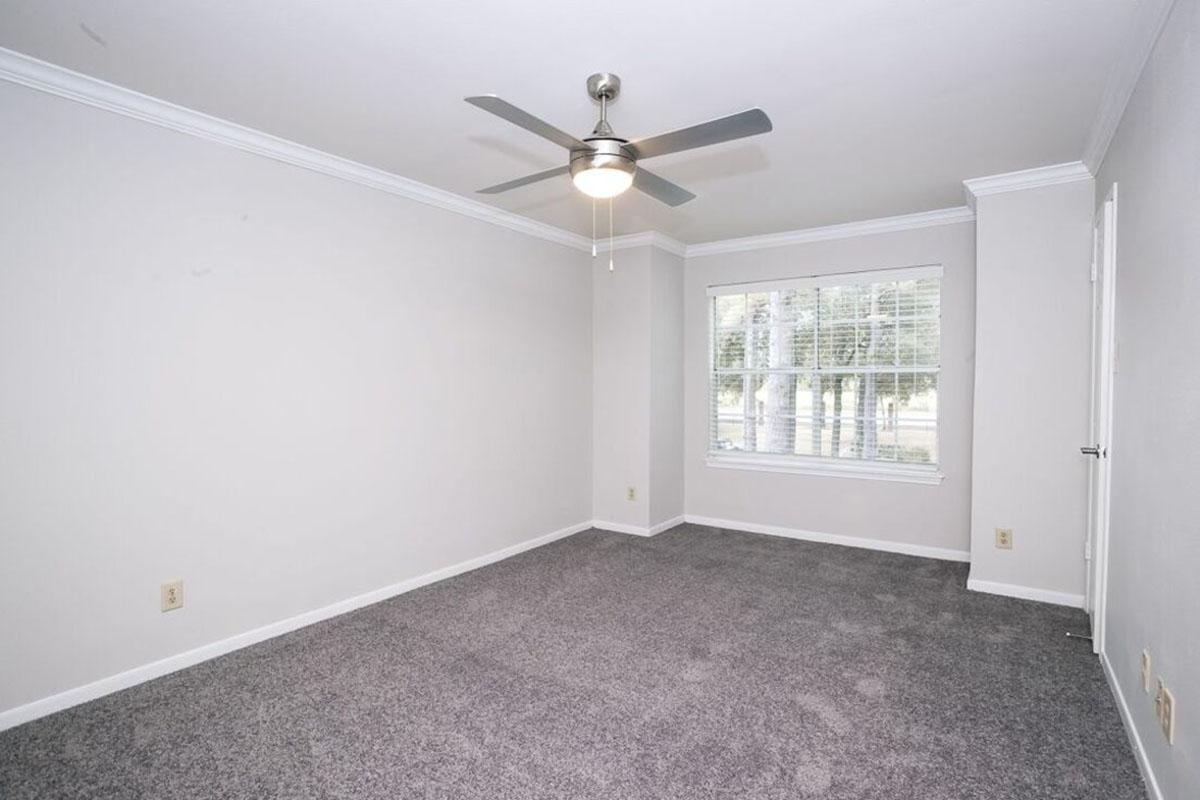 A spacious, empty room with light gray walls and carpet. A ceiling fan is installed in the center, and a large window with multiple panes allows natural light to enter. The room features a simple design with a clean aesthetic, ideal for personal customization.
