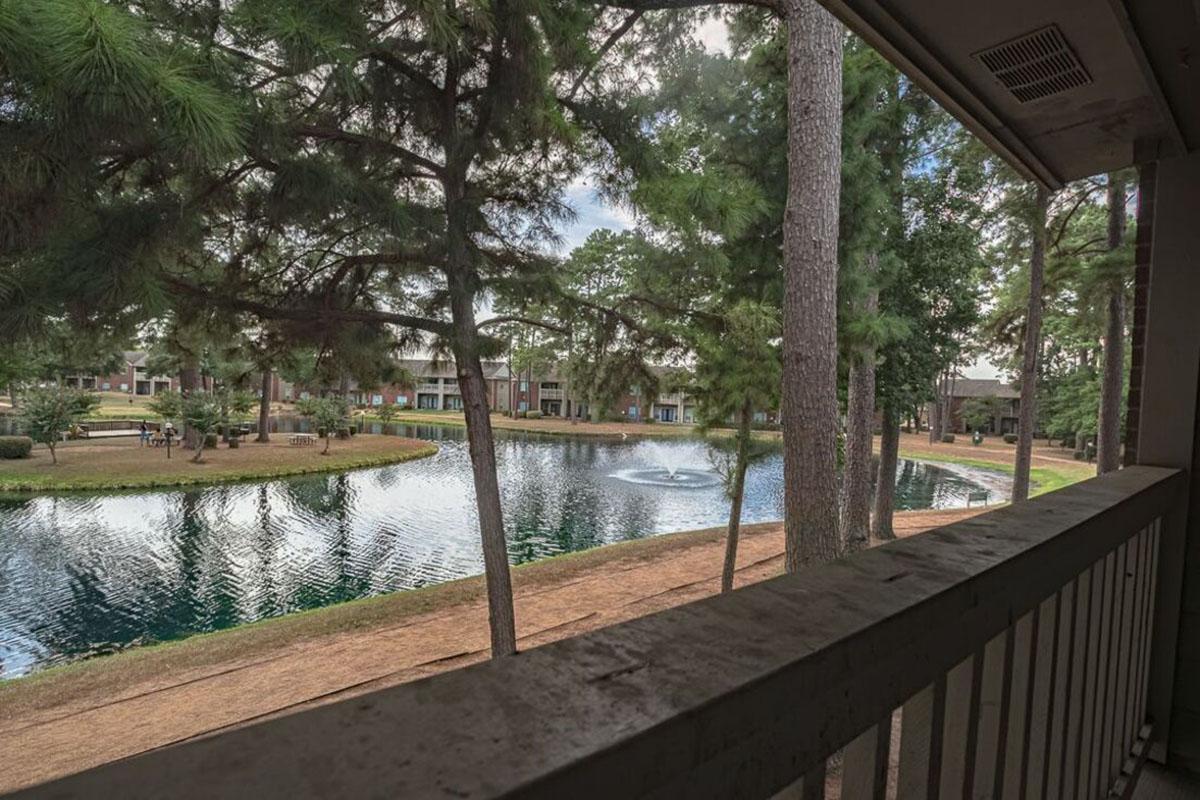 View from a balcony overlooking a serene pond surrounded by tall pine trees. The water reflects the sky and greenery, with a fountain gently spraying in the center. There are manicured lawns and pathways leading around the pond, creating a peaceful outdoor atmosphere.