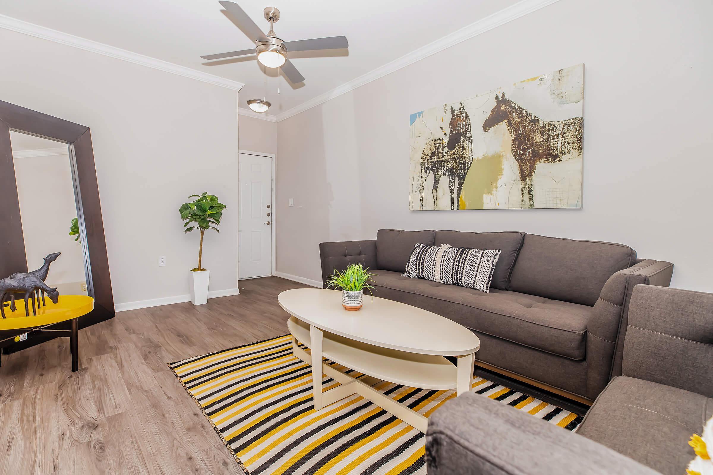 A modern living room featuring a gray couch with black and white patterned pillows, a circular coffee table, and a striped area rug. A large mirror reflects the space, and a painting of horses adorns the wall. A small potted plant is on the table, and a black decorative horse is on a yellow side table.