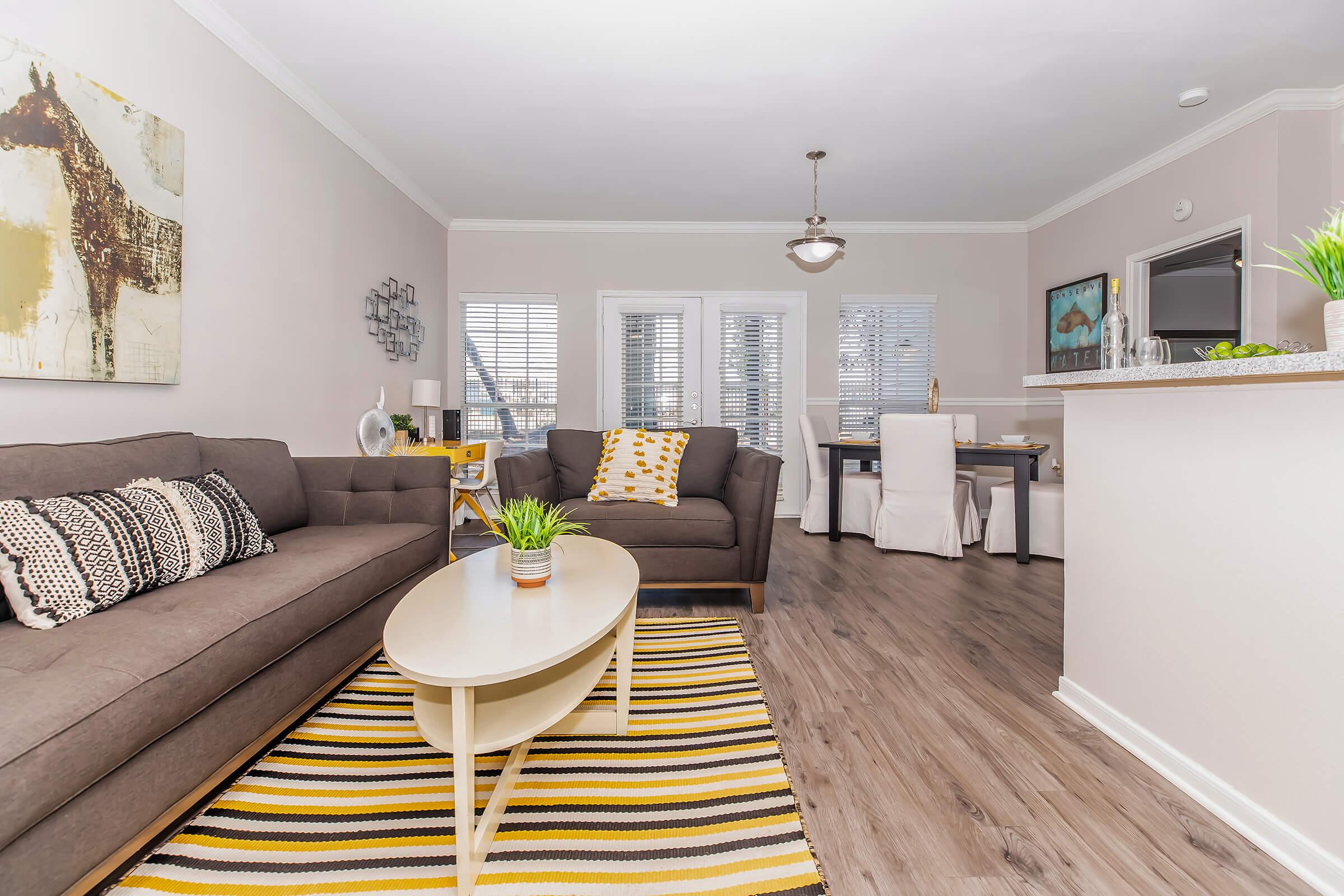 A cozy living room featuring a dark gray sofa and armchair, a round coffee table, and a striped area rug. Large windows allow natural light to flood the space, and a dining area is visible in the background. Decorative plants and artwork add a touch of style to the modern design.