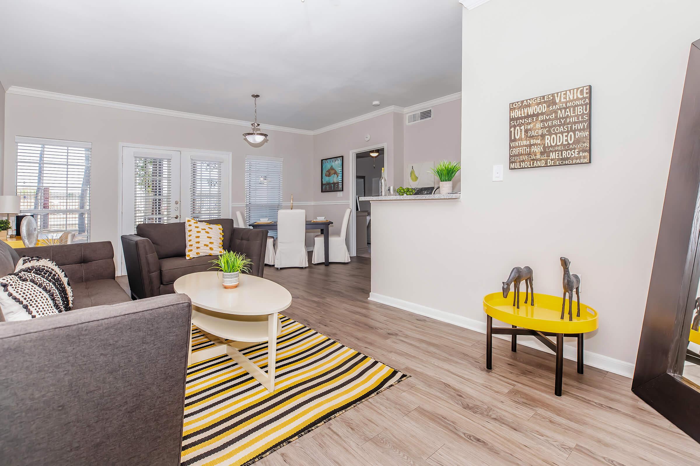 Bright living room featuring a comfortable gray sofa, a circular coffee table, and a striped yellow and black rug. The space is decorated with plants and wall art, with a dining area visible in the background. Large windows let in natural light, enhancing the inviting atmosphere of the modern interior.