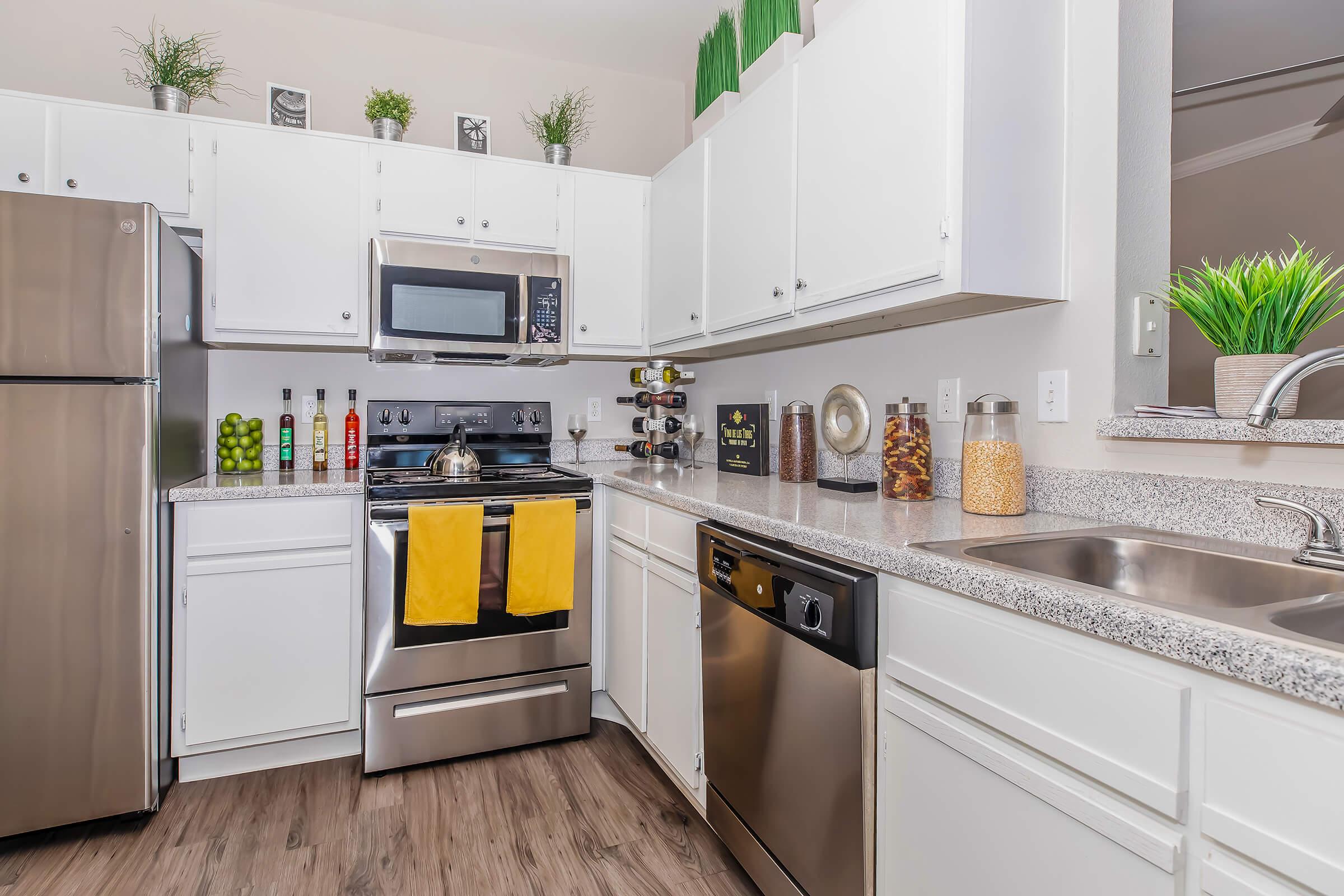 Modern kitchen featuring white cabinetry, stainless steel appliances, and a granite countertop. The kitchen includes an oven, microwave, and dishwasher, with decorative items like green plants and condiments arranged on the counter. A vibrant yellow dish towel hangs from the stove. Wood-like flooring completes the space.
