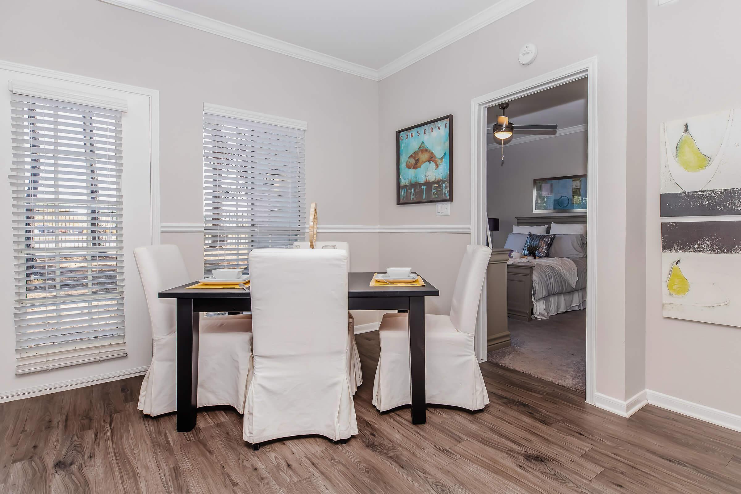 A modern dining area featuring a dark wooden table set with white dishware, surrounded by four white slipcovered chairs. Natural light filters through the blinds on two windows, with neutral-colored walls and stylish artwork adding a touch of decor. A doorway leads to a cozy living space in the background.