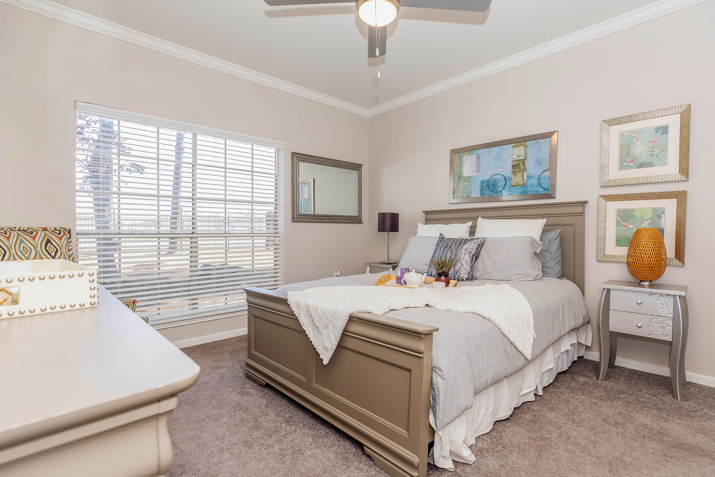 A cozy bedroom featuring a light gray bed with decorative pillows, a bedside table, and a small lamp. Natural light streams through a window with blinds, illuminating the room. The walls are adorned with colorful artwork, and a mirror reflects the soft, inviting atmosphere.