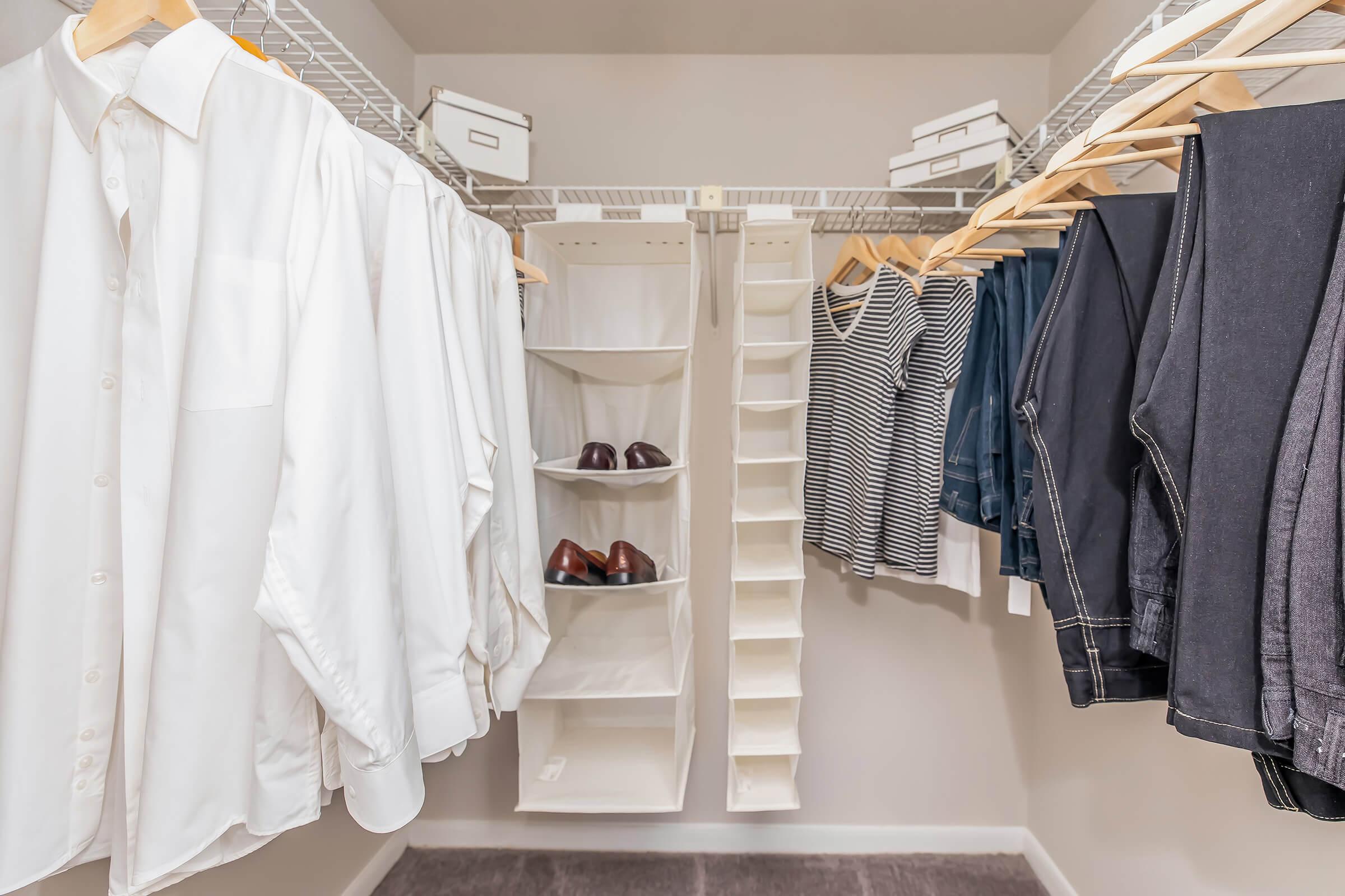 A well-organized closet featuring neatly hung white shirts on wooden hangers, a few pairs of dark pants, and a striped tank top. There are also brown shoes on a shelf and storage boxes above. The space is well-lit and clean, demonstrating a minimalistic and tidy arrangement.