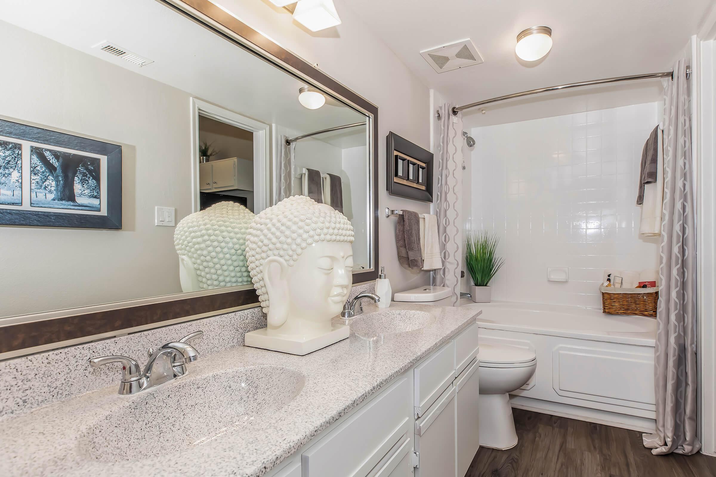 Modern bathroom featuring a double sink with a mirrored wall, a decorative Buddha head sculpture, and a bathtub with a shower. Neutral tones create a calming atmosphere, complemented by a small plant and a framed picture on the wall.