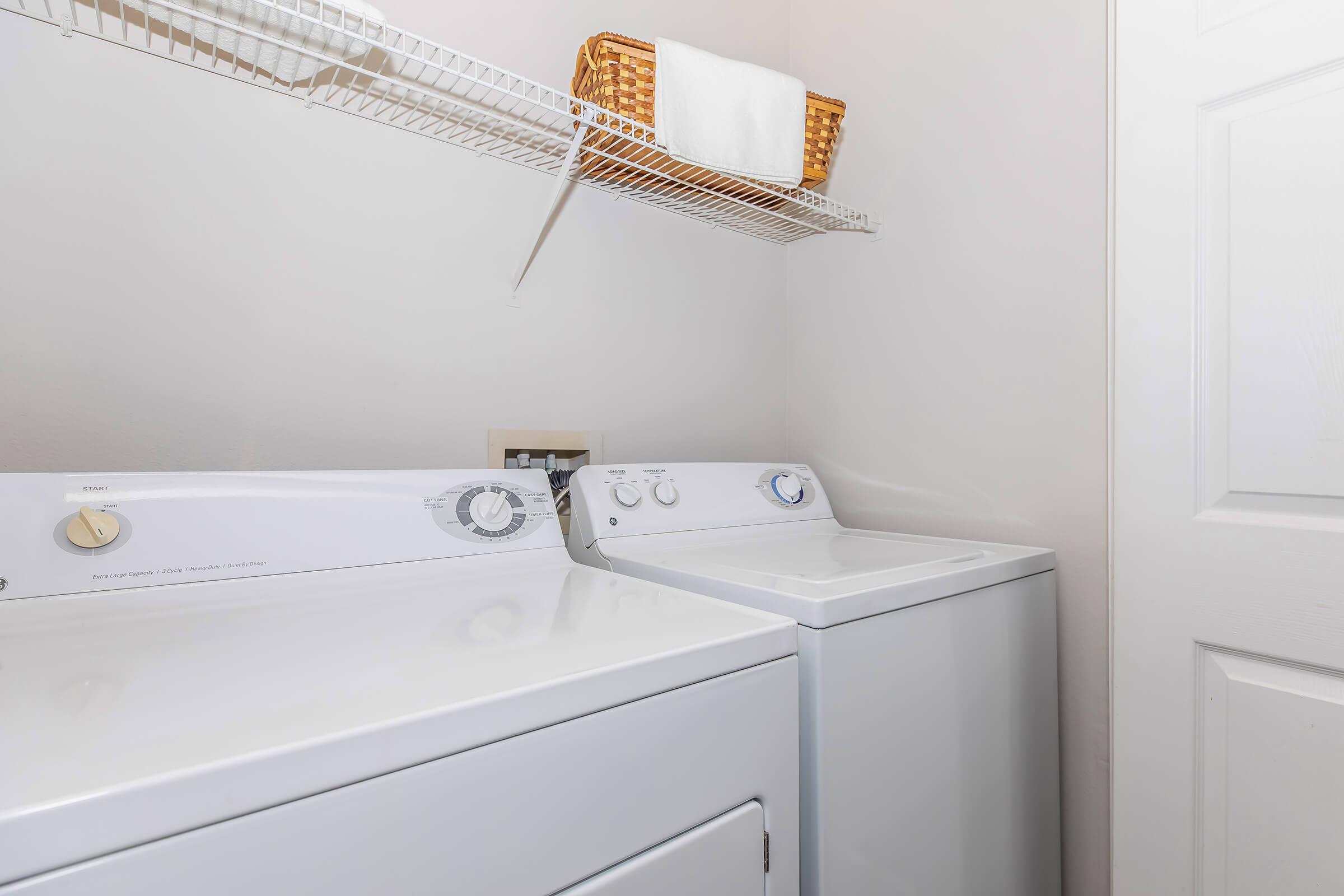 A clean and organized laundry room featuring a white washing machine and dryer side by side, with a wire shelf above holding a basket and a folded towel. The walls are light-colored, enhancing the bright, airy feel of the space.