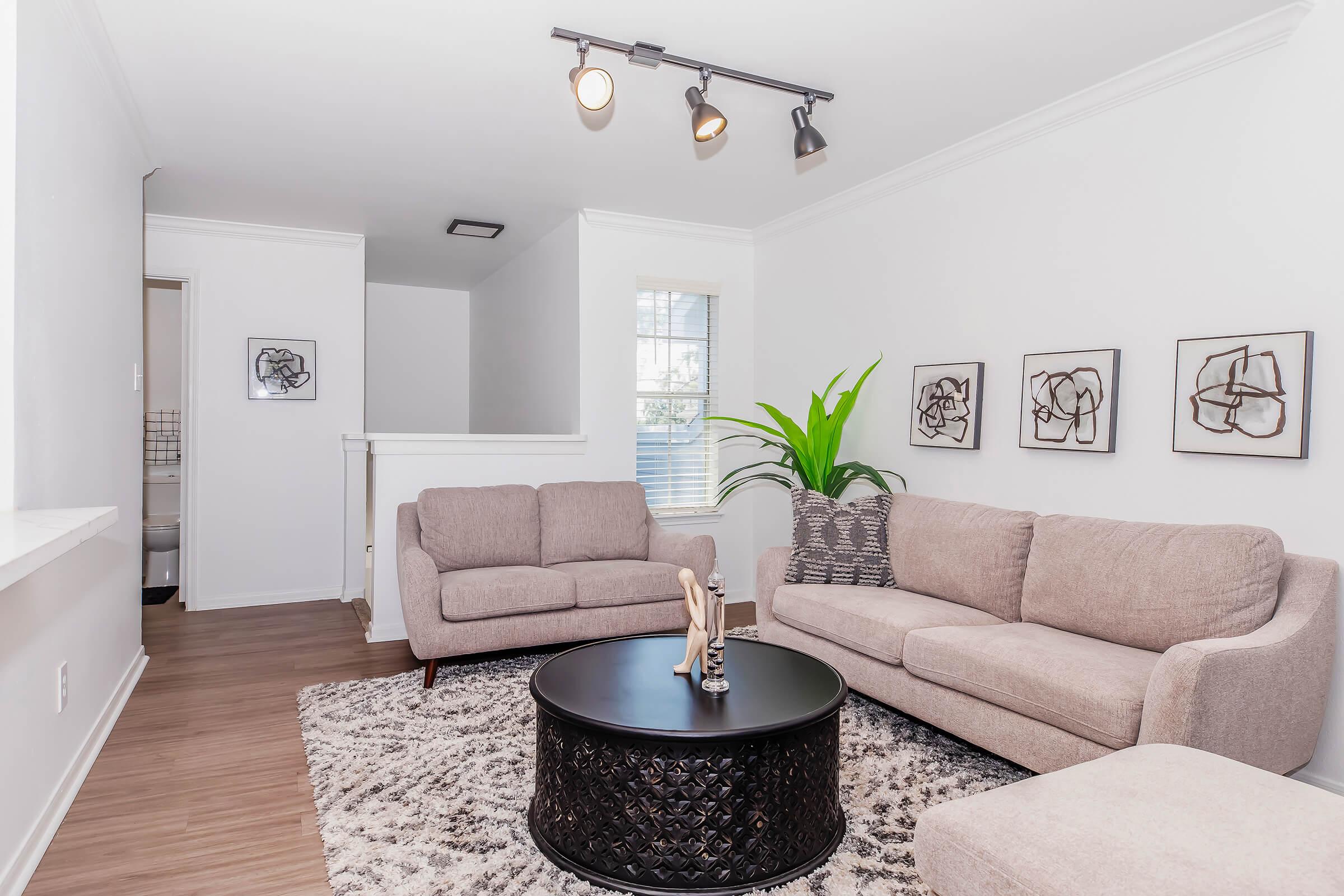 A modern living room featuring two light-colored sofas, a round black coffee table, and decorative wall art. A large green plant adds a touch of nature, while warm lighting illuminates the space. The room has a cozy atmosphere with a soft area rug on a hardwood floor.