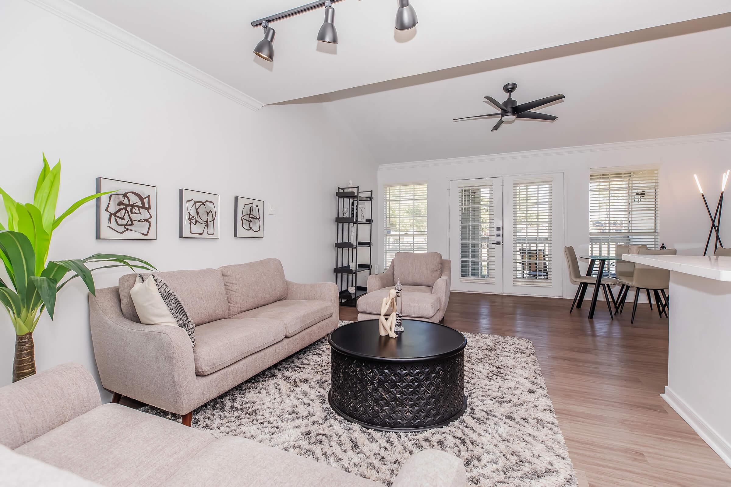 A cozy living room featuring two light gray sofas, a round black coffee table, and decorative wall art. There is a potted plant in one corner and a black shelving unit nearby. The space is brightened by natural light from large windows with blinds, leading to a dining area in the background.