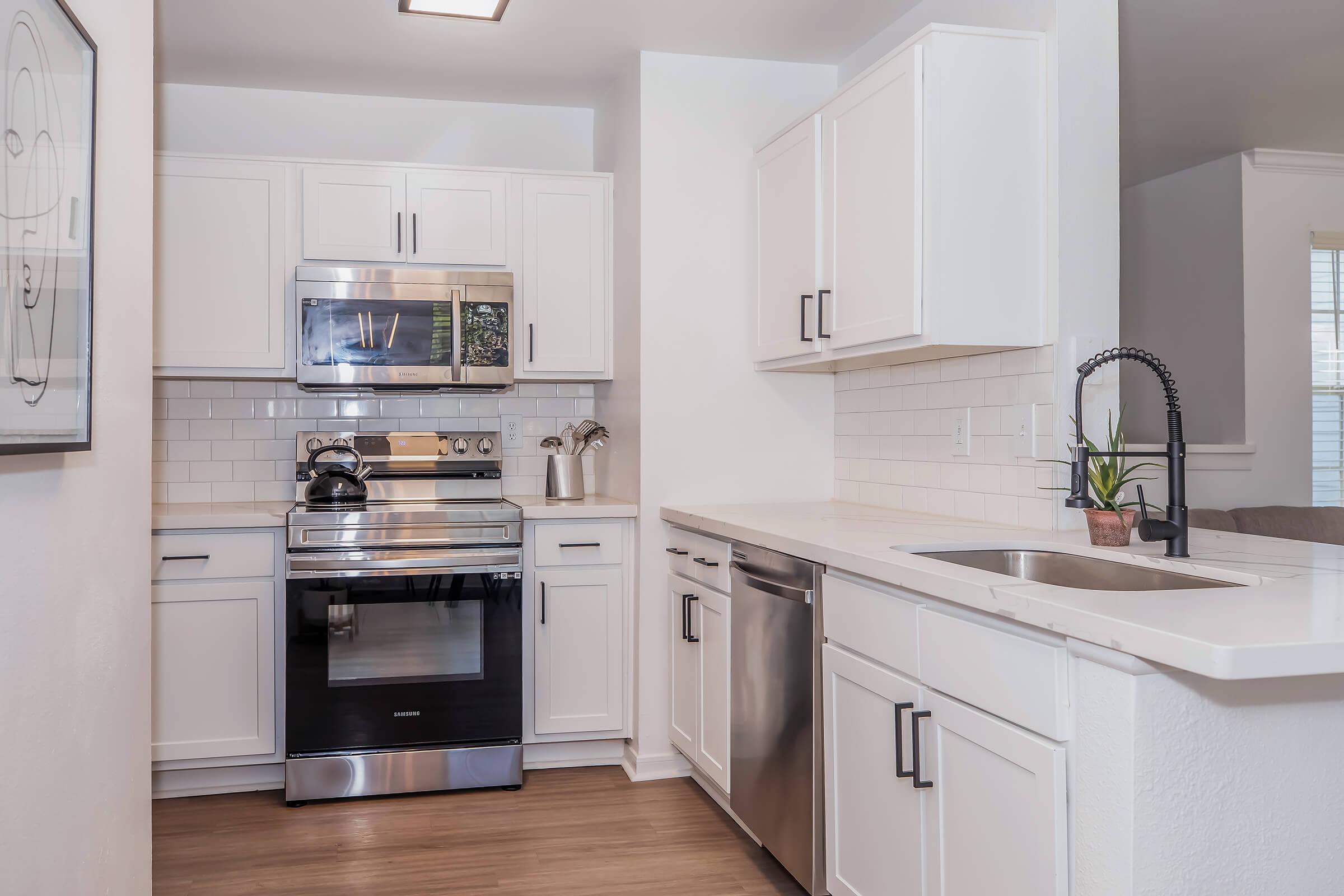 Modern kitchen featuring white cabinets, stainless steel appliances including a microwave, oven, and dishwasher, a light countertop, and a sink with a sleek faucet. The backsplash consists of white subway tiles, and there is a small plant on the countertop. Natural light comes from a nearby window.