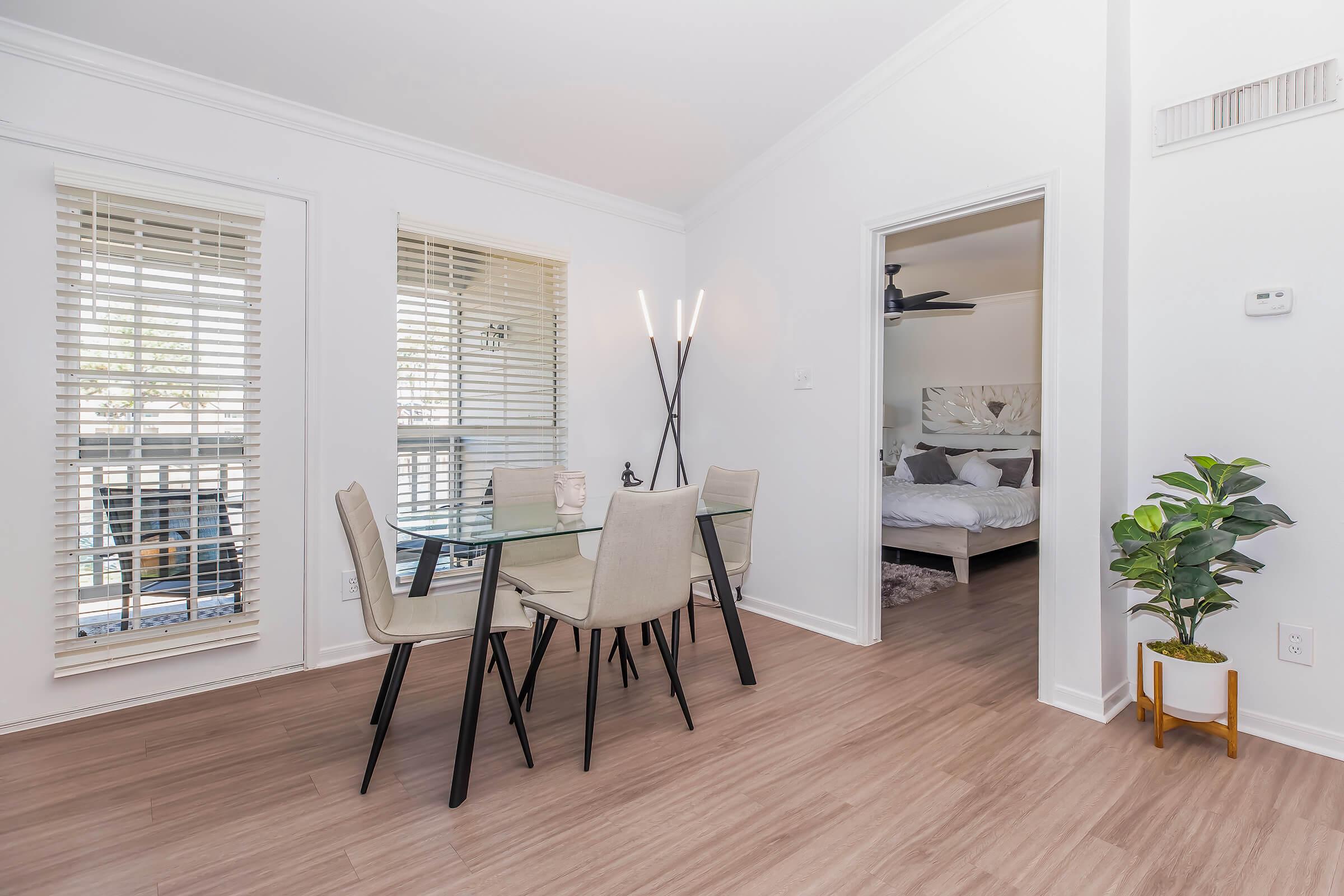 A modern dining area featuring a glass table with four light-colored chairs, surrounded by bright natural light from windows with horizontal blinds. A potted plant adds a touch of greenery, and a doorway leads to a cozy bedroom with a bed visible in the background. The floor is light wood laminate.