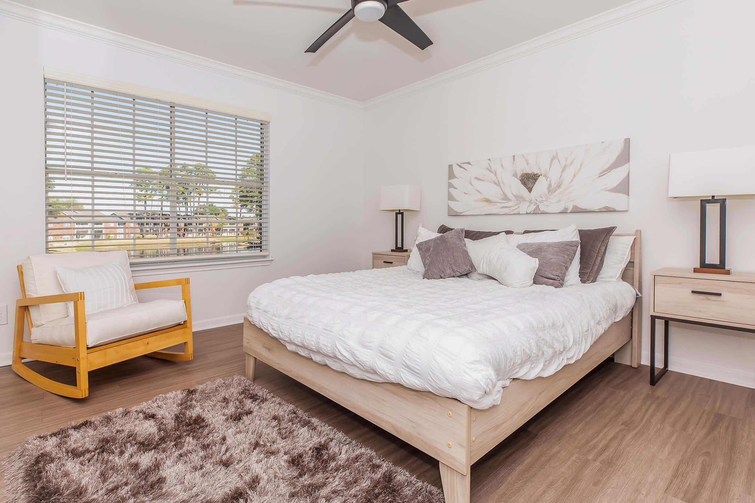 A modern bedroom featuring a comfortable bed with decorative pillows, a cozy rocking chair, and wooden nightstands with lamps. Natural light filters through a window with blinds, illuminating the space and highlighting the neutral color palette and soft textures. A plush area rug adds warmth to the room.