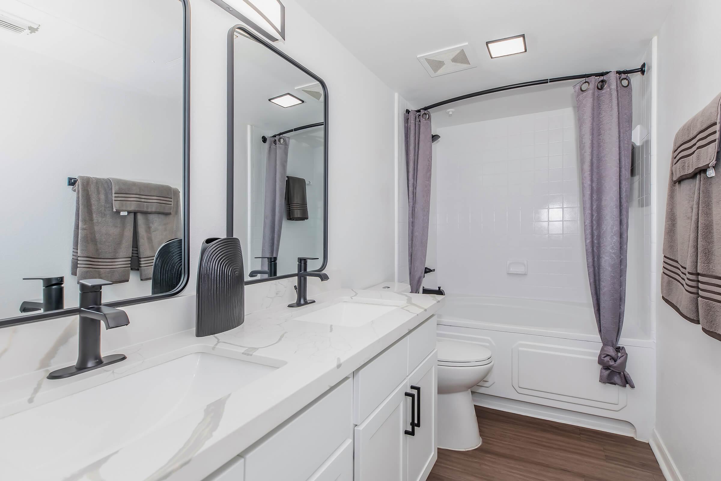 A modern bathroom featuring dual sinks with black faucet fixtures, large mirrors, a bathtub with a shower curtain, and stylish gray towels hanging. The walls are painted white, and the flooring is a warm wood tone, creating a clean and contemporary aesthetic.