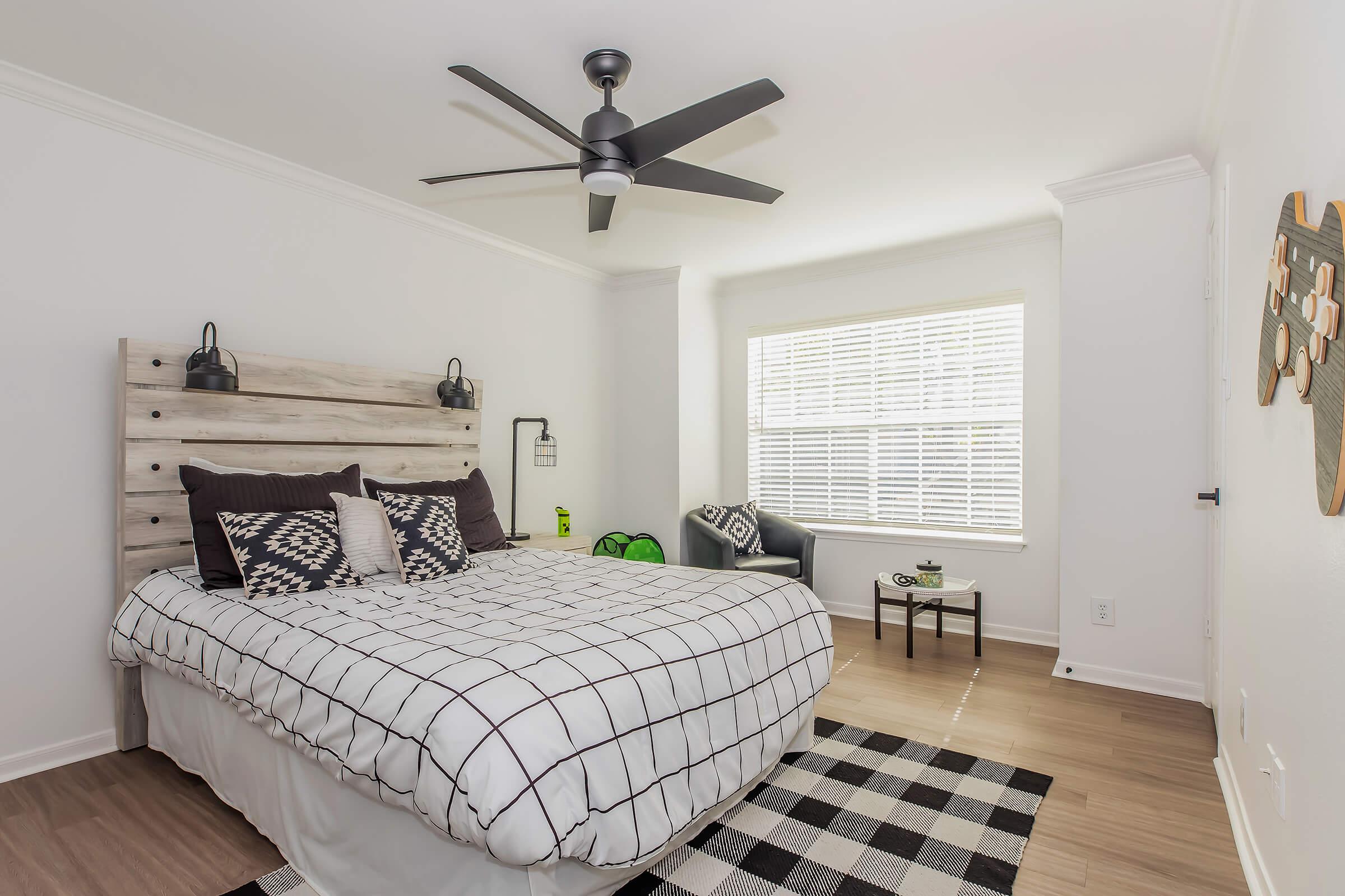 A modern bedroom featuring a bed with a plaid duvet and decorative pillows, a wooden headboard, a ceiling fan, and large windows with blinds. A cozy seating area includes a chair and a small round table, with decorative accents on the wall and a green plant in the corner.