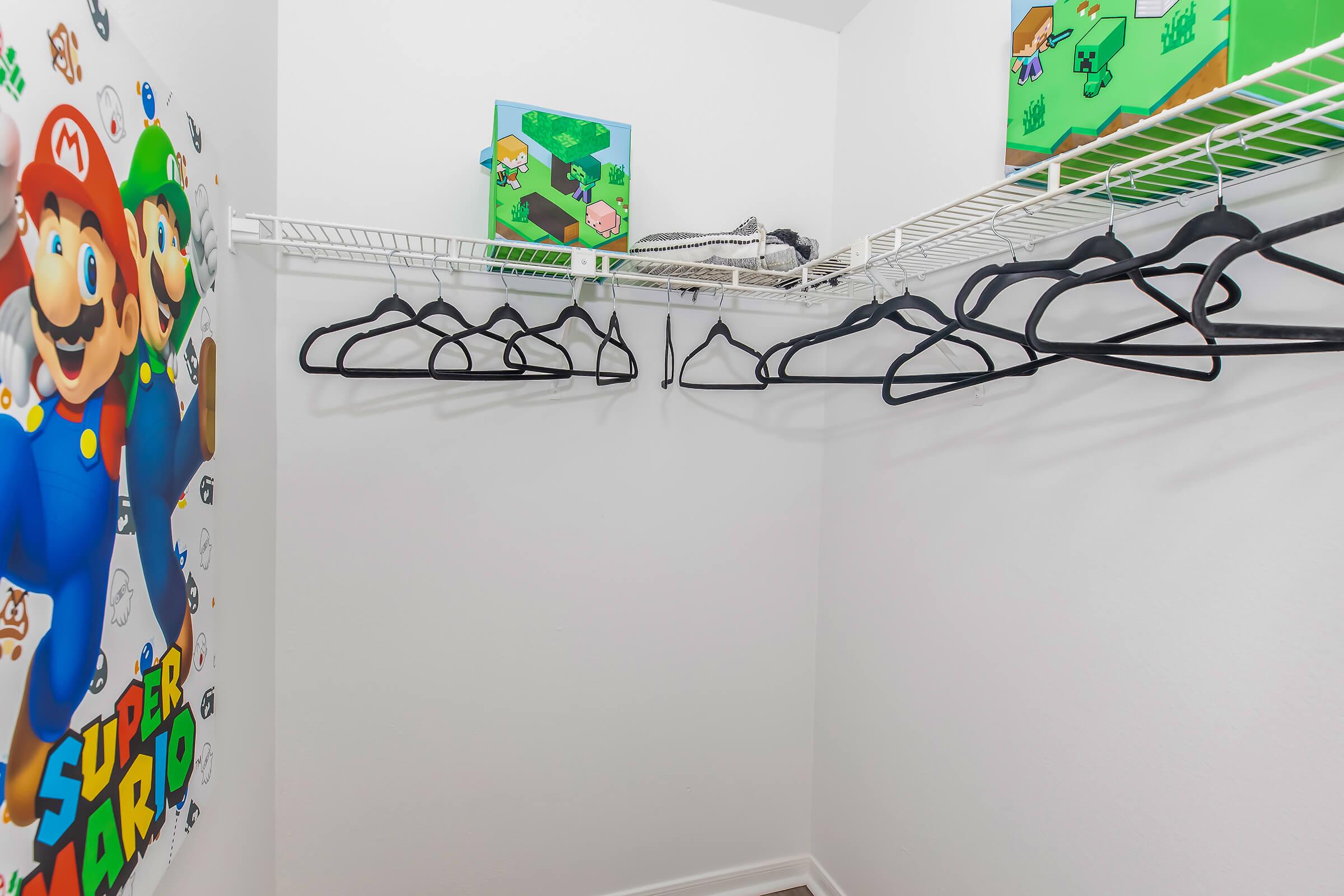A closet with white walls and a wire shelf. The shelf contains a few items, and there are several black hangers hanging below it. One wall features a colorful mural of Mario from the Super Mario video game series, adding a playful touch to the space.