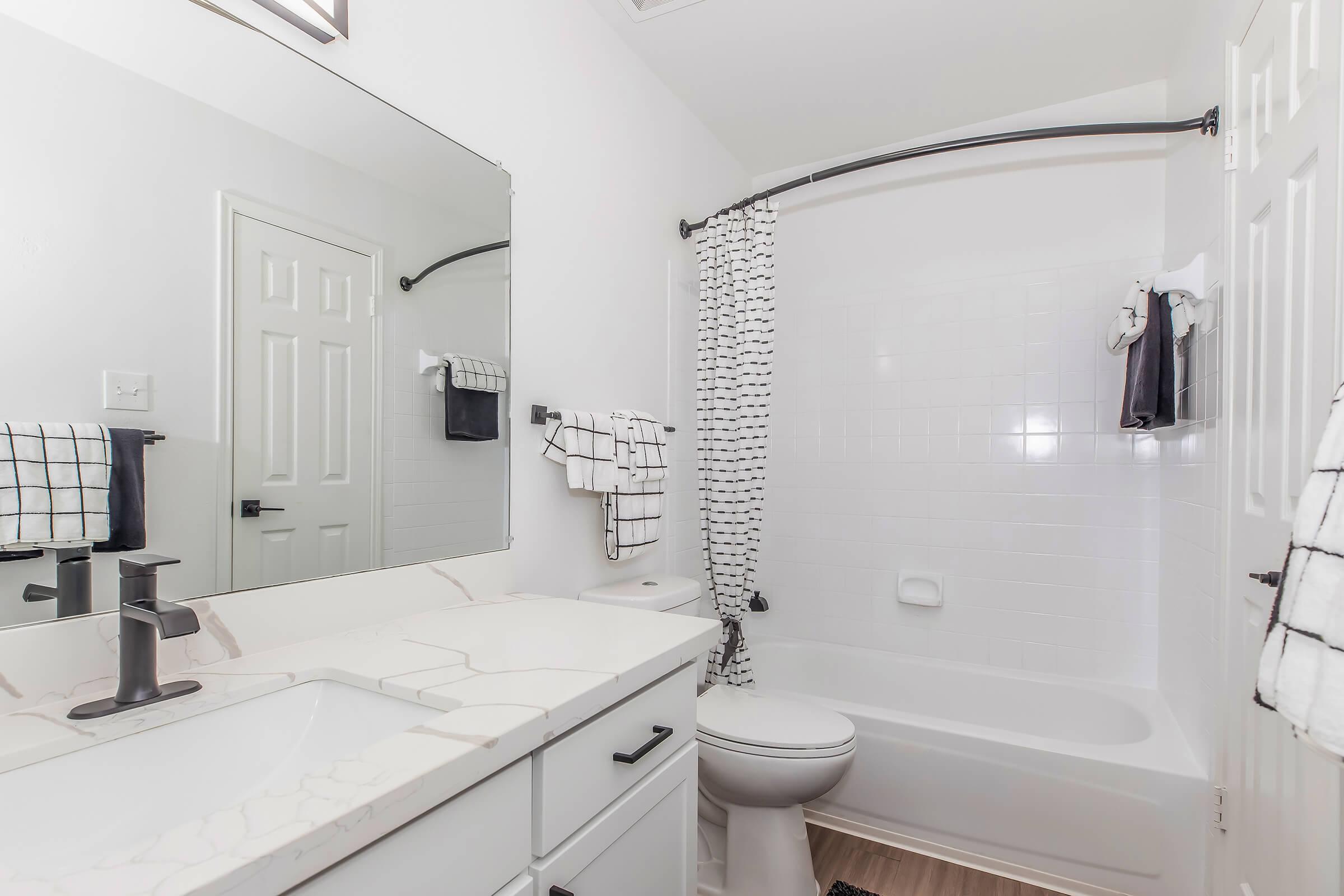 Modern bathroom featuring a white tiled tub and shower, a sleek sink with a marble countertop, and large mirror. Black and white checkered towels hang on a rack, with a black shower curtain adding contrast. Bright, clean aesthetic with minimal decorations.