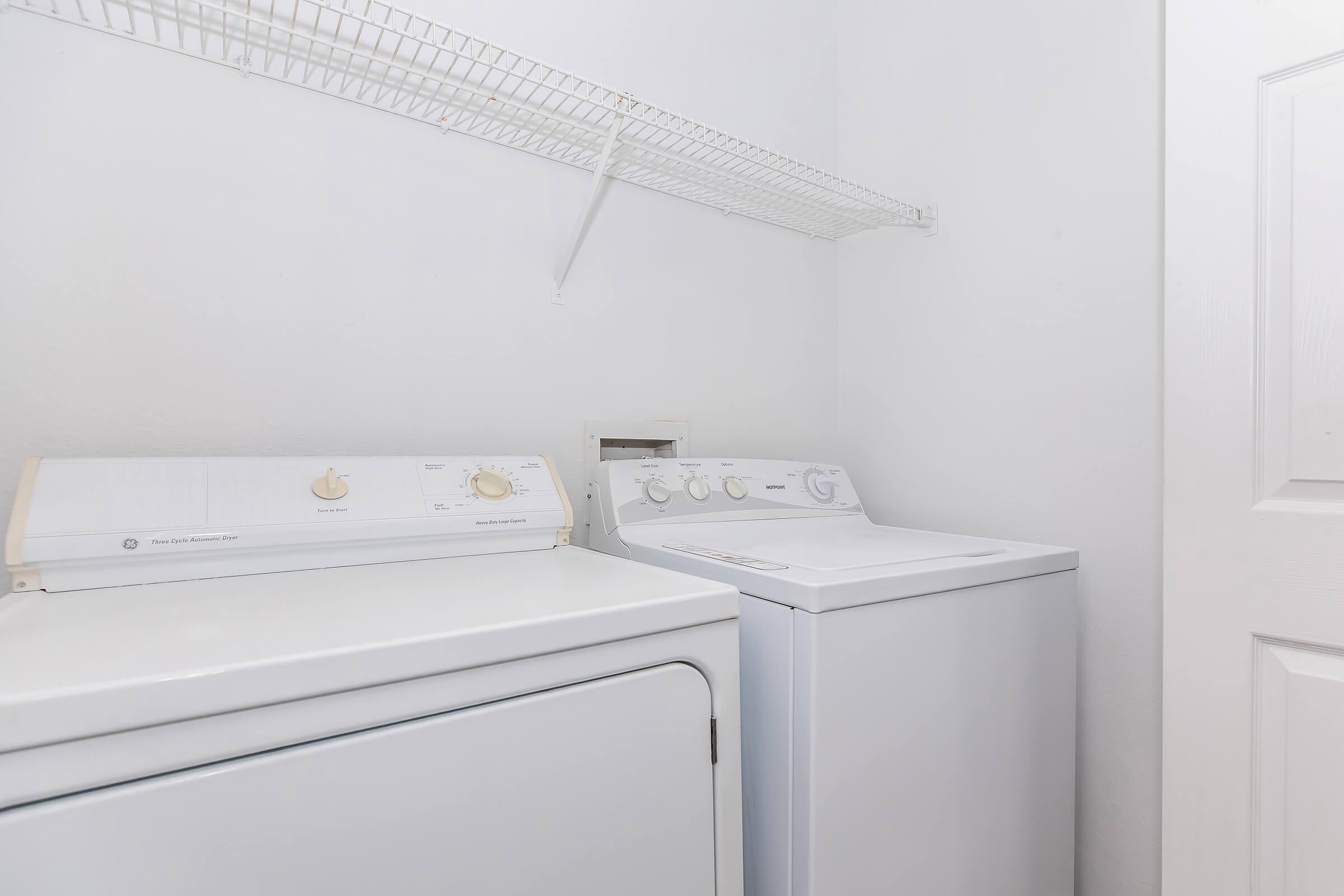 A clean laundry room featuring a stacked washer and dryer, both in white. The appliances are located against a light-colored wall, with a wire shelf mounted above for storage. The space is well-lit and organized, creating a functional laundry area.