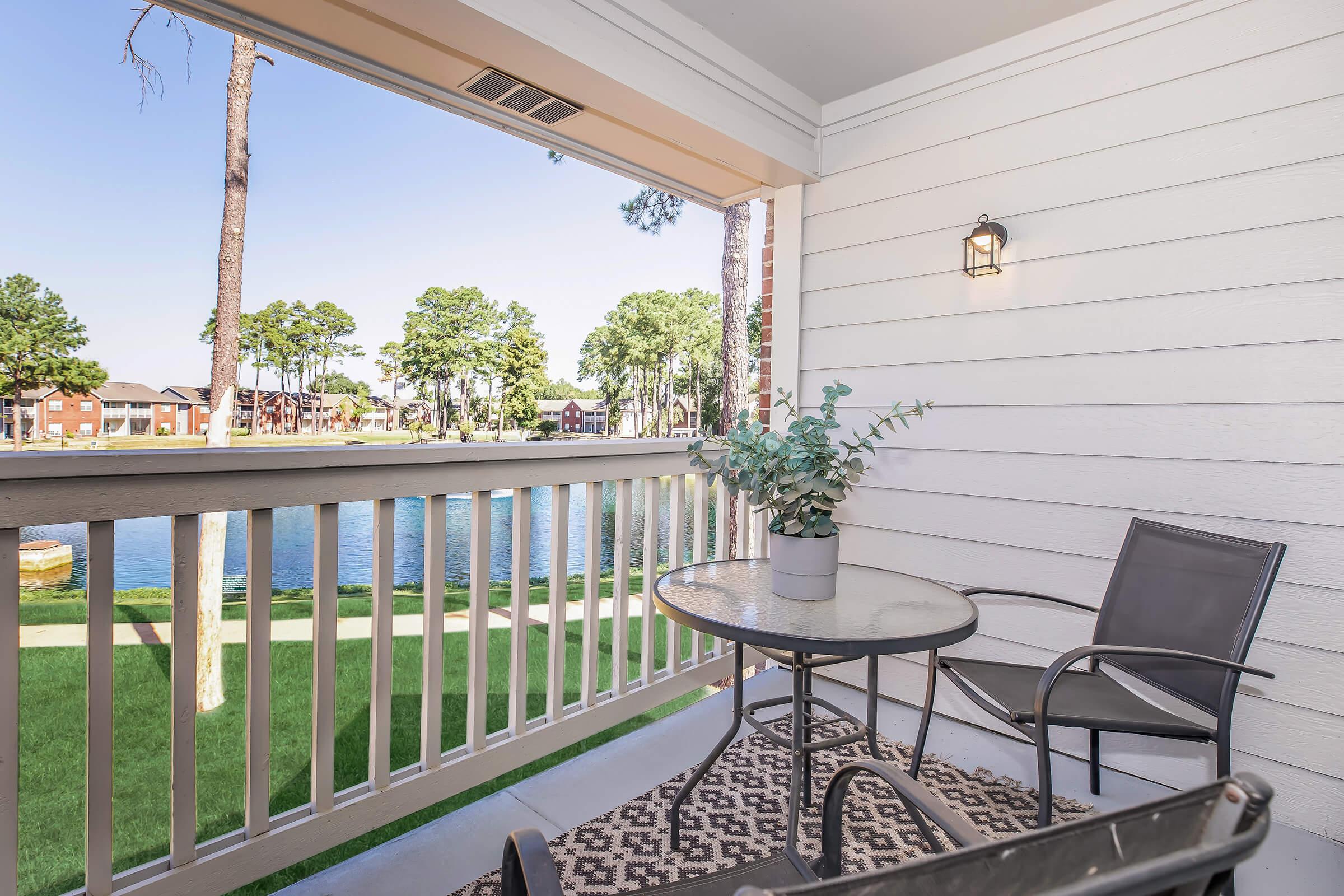 A cozy balcony with a round glass table and a single chair, featuring a potted plant. The view includes a serene body of water and trees in the background, creating a peaceful outdoor atmosphere. The balcony has a light fixture and is surrounded by wooden railings.