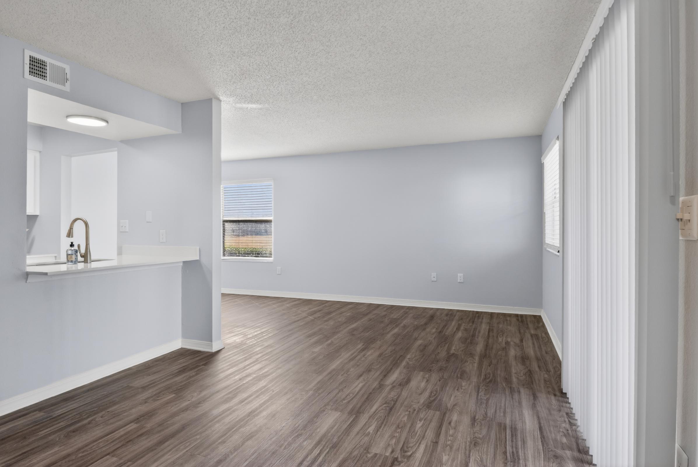 A spacious, empty living area with light-colored walls and wood-style flooring. There is a small kitchen bar area with a sink visible on the left. Natural light enters through a window, and vertical blinds are present. The room appears freshly painted and ready for furnishings.