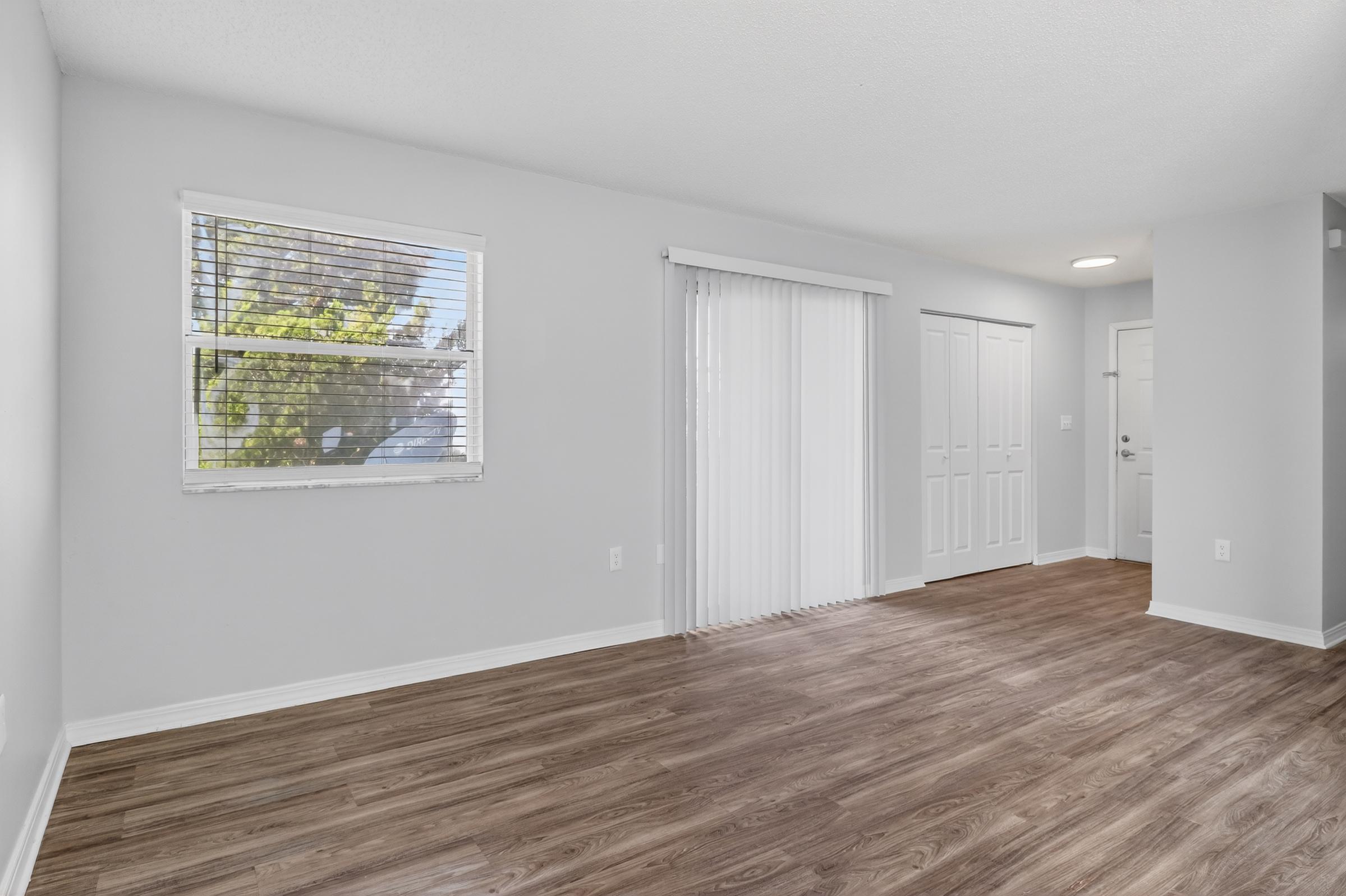 A bright, empty living room featuring light gray walls, a window with blinds, and a sliding glass door that leads outside. The floor is covered in light wood laminate. Two white closet doors are visible on the right side, and there's a door that likely leads to an entryway.