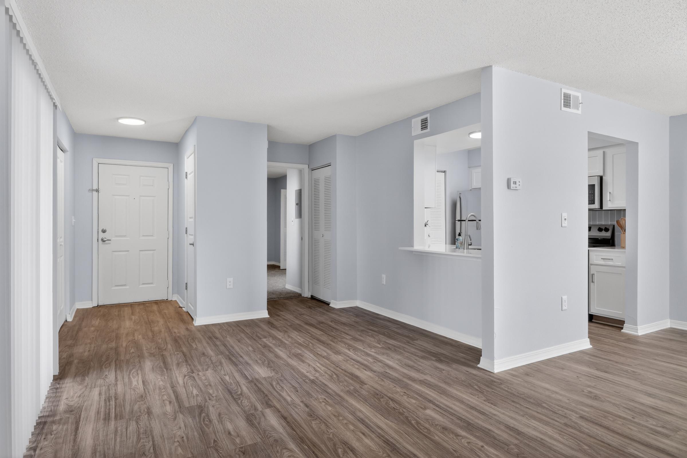 Bright, modern living space featuring light blue walls and hardwood flooring. The layout includes an entrance door, doorways leading to other rooms, and a kitchen area visible through an open pass-through. Natural light enhances the airy feel of the room.