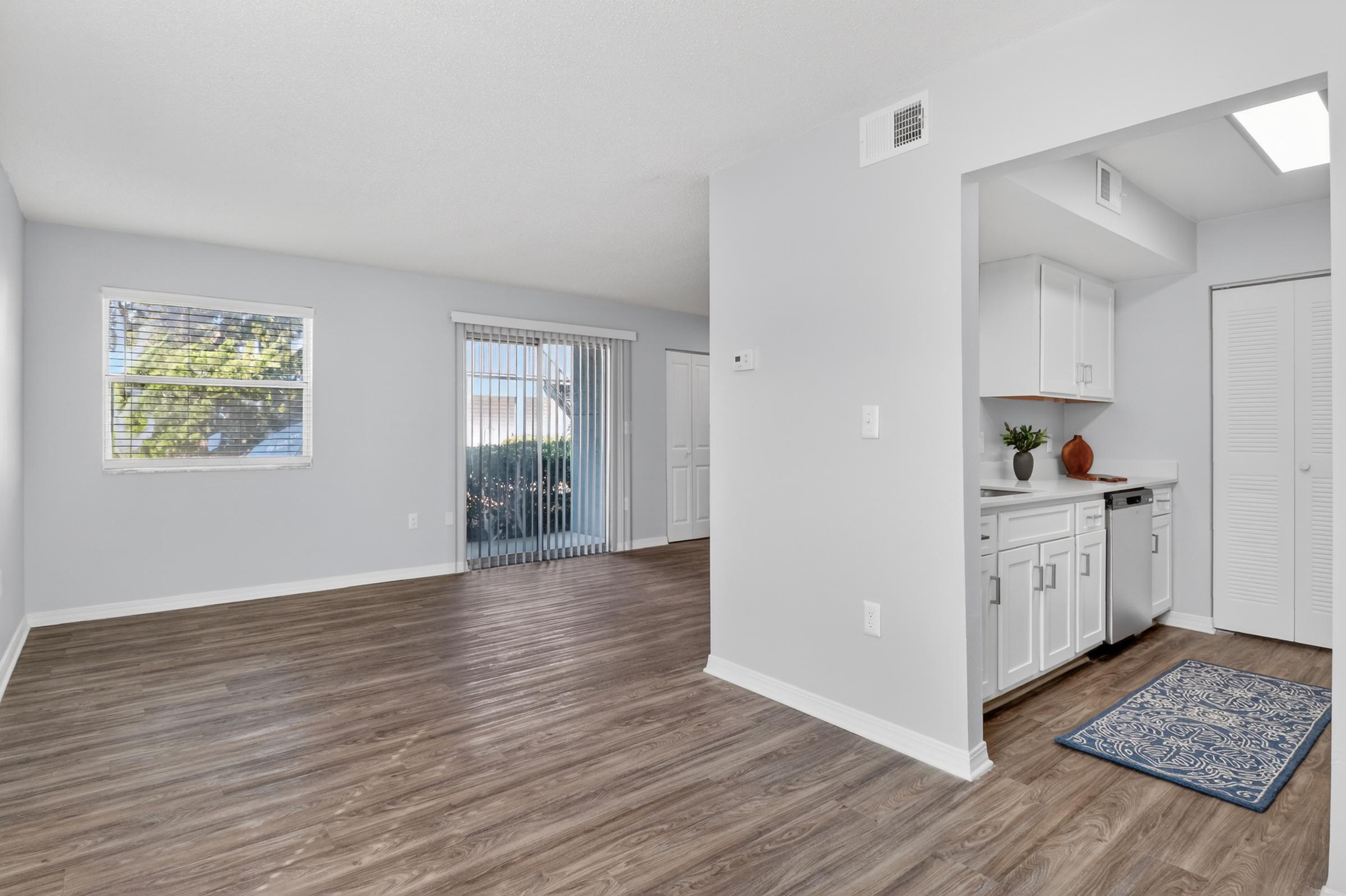 A light and airy living space featuring wooden flooring, neutral-colored walls, and large windows. The room includes a partially visible kitchen area with modern appliances and a decorative rug. Sliding glass doors lead to an outdoor space, creating a bright and inviting atmosphere.