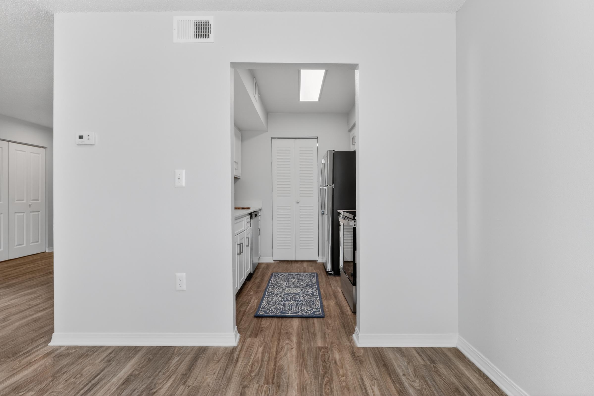 A view of a modern kitchen with white cabinetry and black appliances. The space features wooden flooring, a rug in the center, and a bright overhead light. A doorway leads to a separate area, with doors visible on the left side. The walls are painted in a light color, creating a clean and open atmosphere.