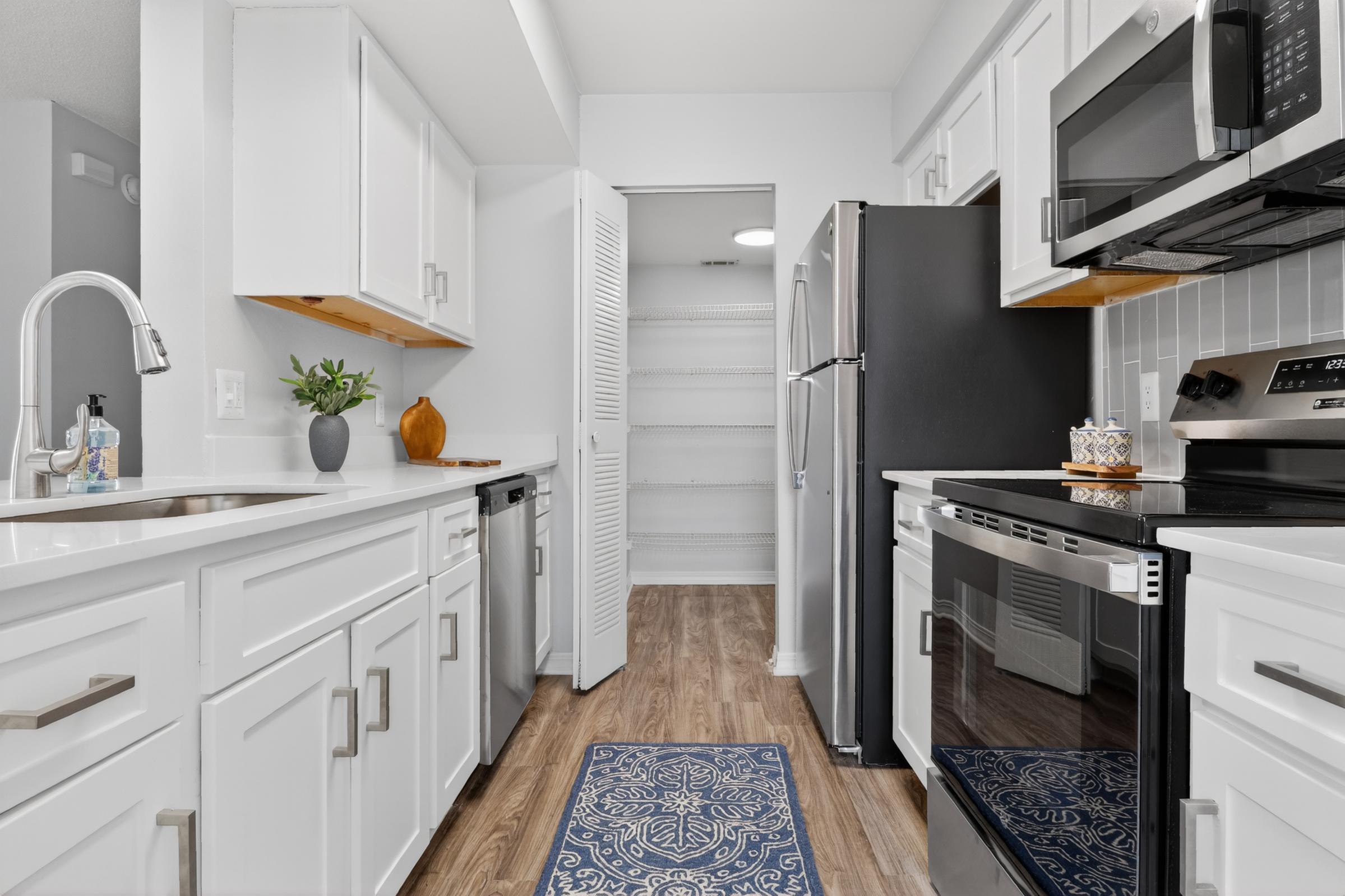 A modern kitchen featuring white cabinetry, stainless steel appliances including a refrigerator and microwave, and a dark oven. There's a decorative rug on the wooden floor, a small plant on the counter, and a pantry area with closed doors in the background. The space is brightly lit and stylishly designed.