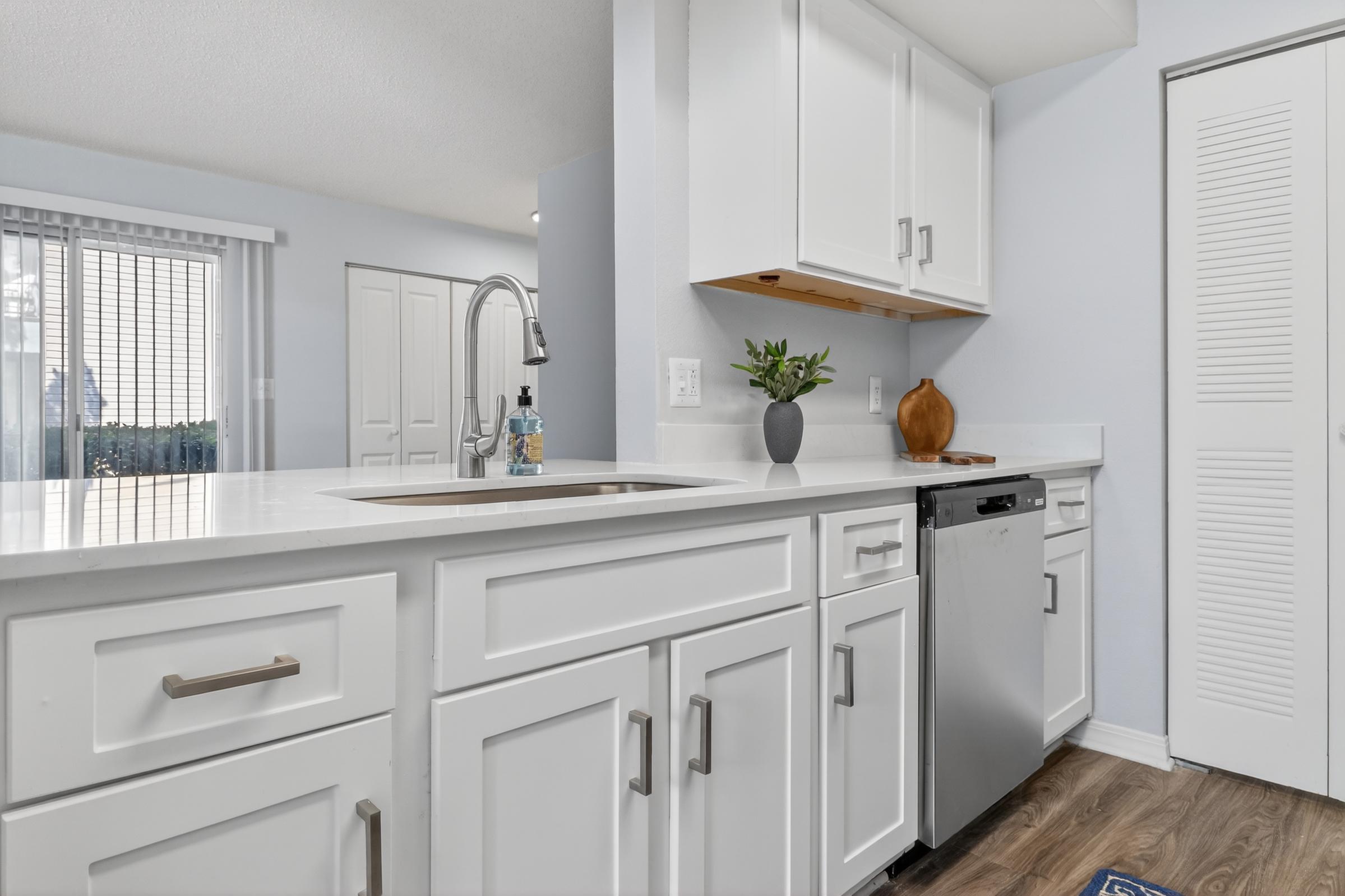 Modern kitchen with white cabinets and a sleek countertop. Features include a stainless steel dishwasher, a sink with a decorative faucet, and a small potted plant on the counter. Natural light filters in through a window with vertical blinds, enhancing the clean and bright atmosphere.